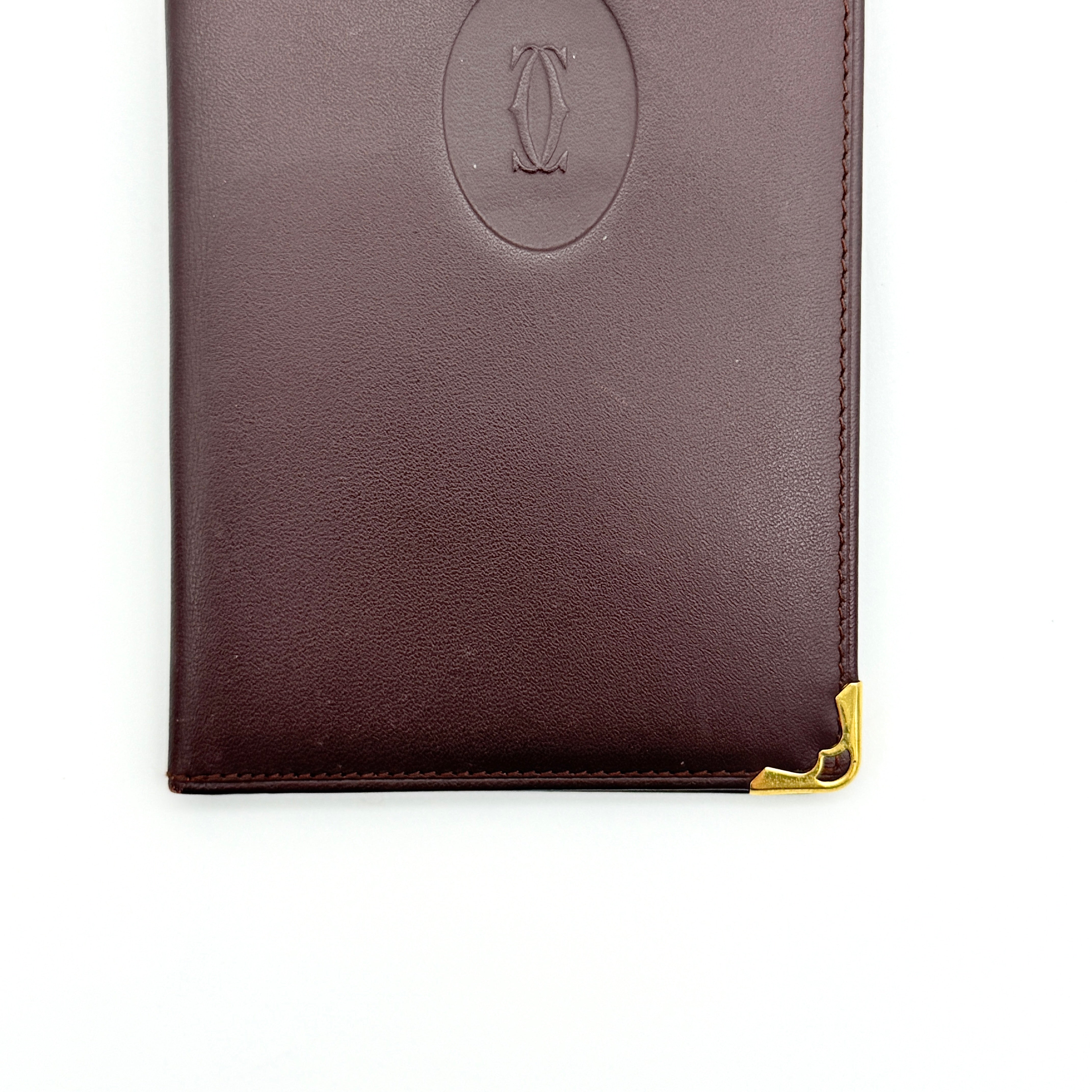 Cartier Must de Cartier bifold long wallet in burgundy leather with gold-tone corner accents, shown closed against a white background.