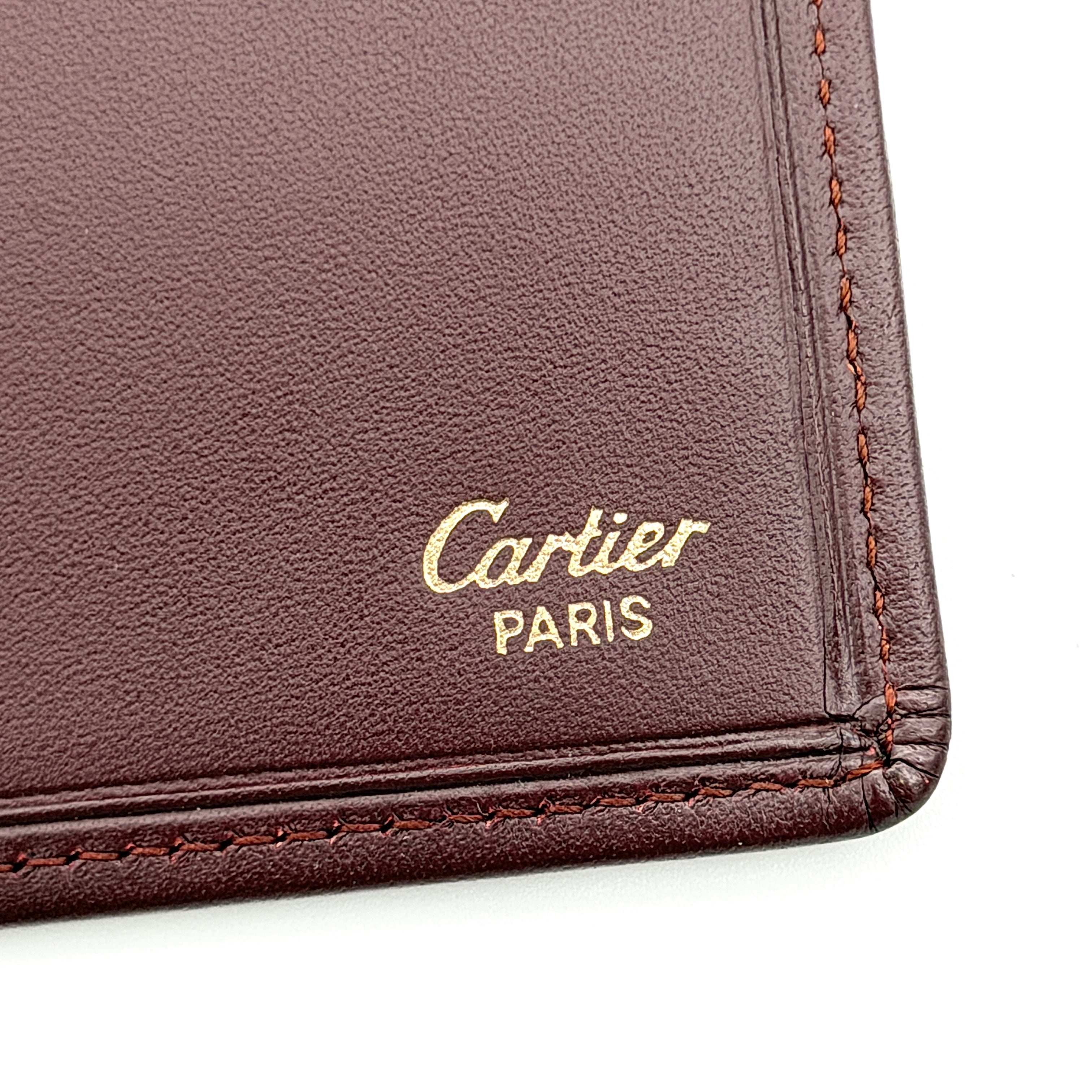 Cartier Must de Cartier bifold long wallet in burgundy leather with gold-tone corner accents, shown closed against a white background.