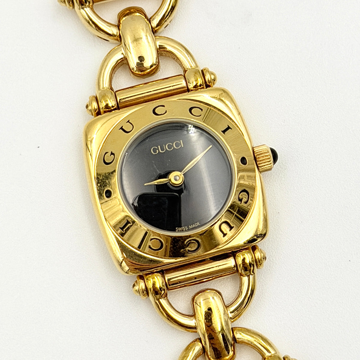 Gucci 6400L gold-tone bracelet watch with a black dial and distinctive horsebit links, showcased on a clear stand.