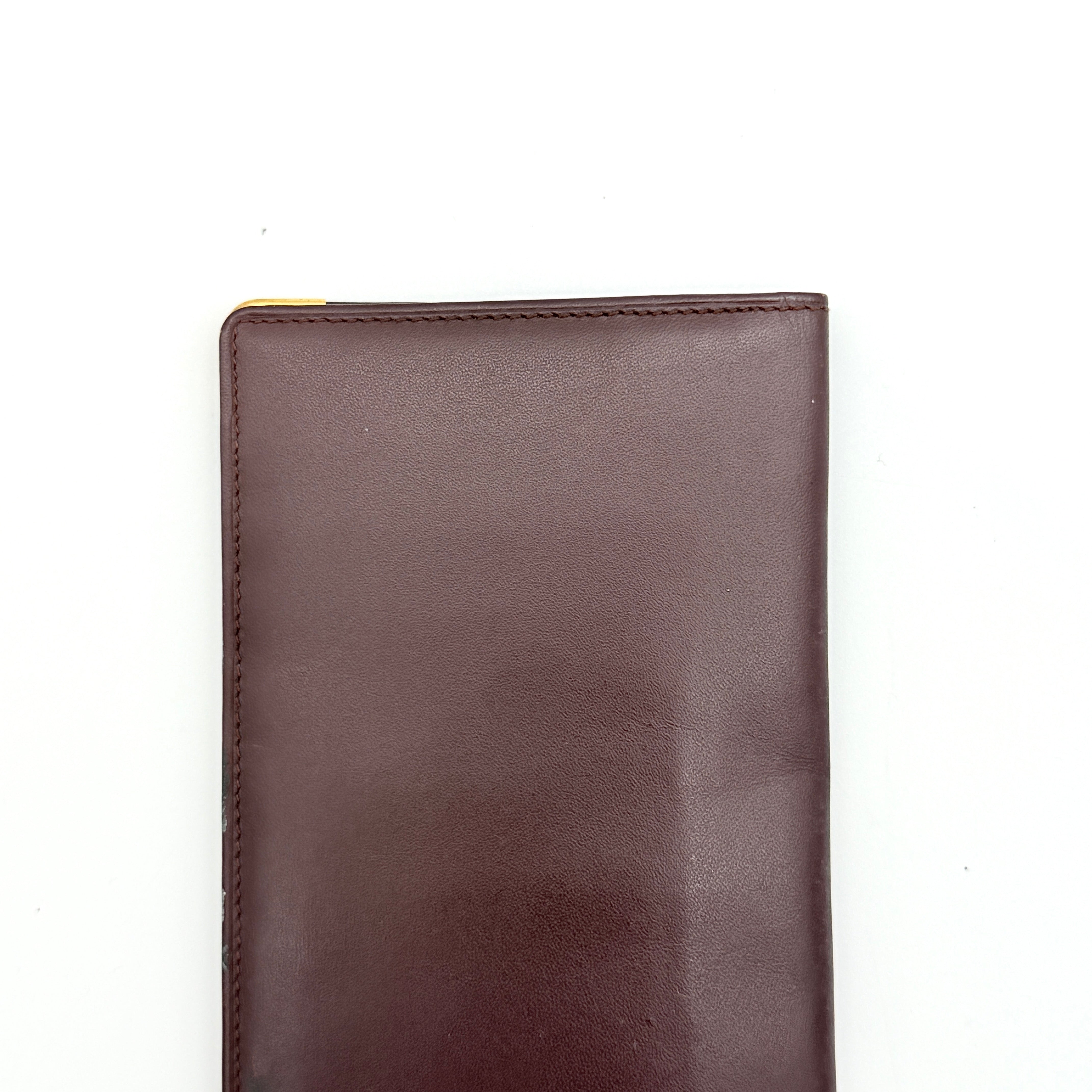 Cartier Must de Cartier bifold long wallet in burgundy leather with gold-tone corner accents, shown closed against a white background.