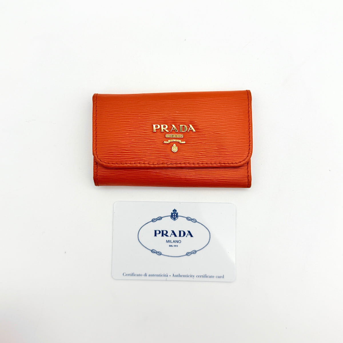 Prada Saffiano leather key case in orange with gold-toned hardware, Prada logo, and authenticity card displayed.