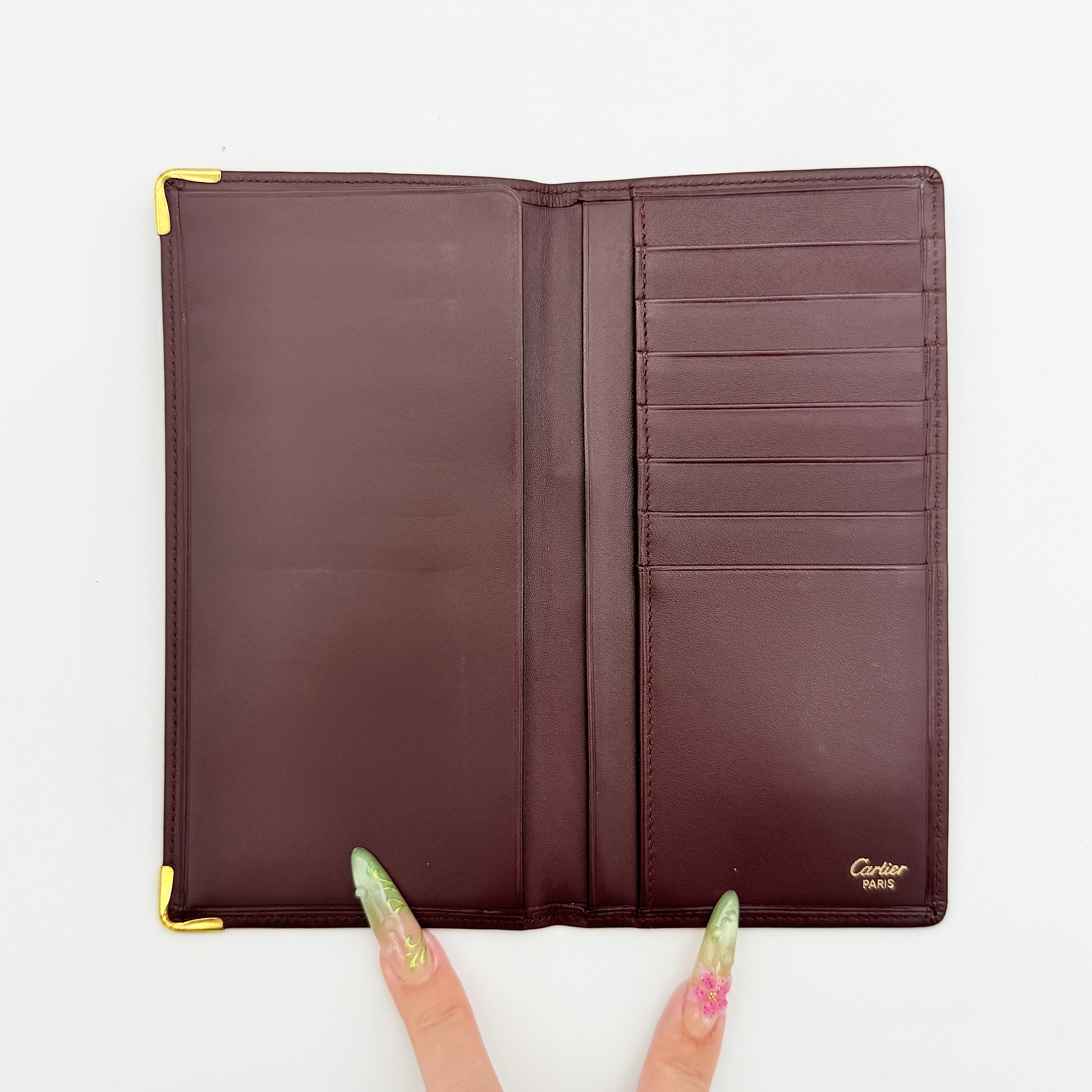 Cartier Must de Cartier bifold long wallet in burgundy leather with gold-tone corner accents, shown closed against a white background.