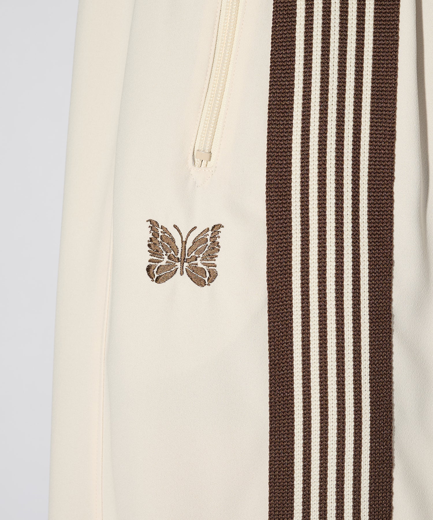 Needles H.D. Track Pant in cream with brown side stripes and embroidered butterfly logo, shown from the side in soft studio lighting.