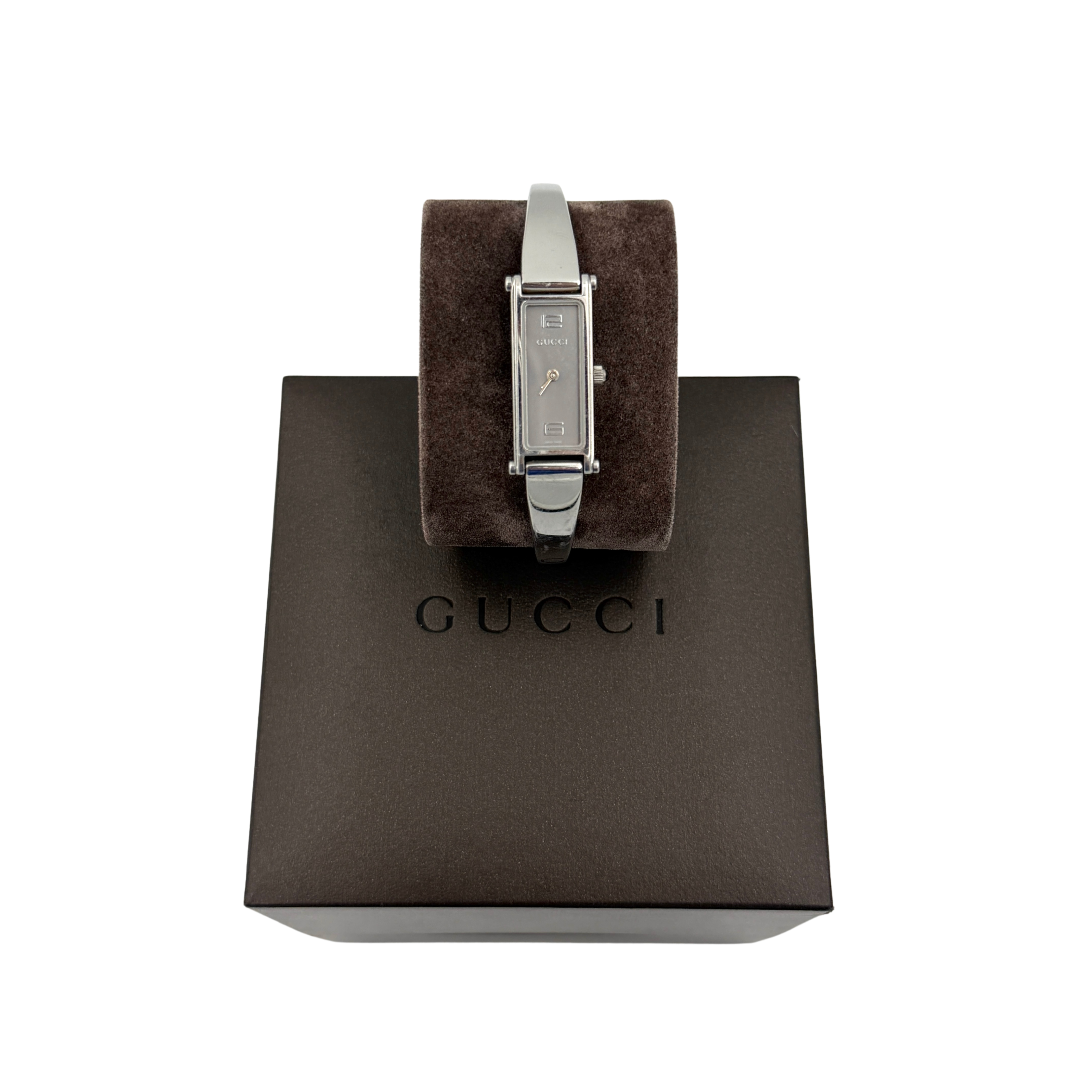 The Gucci 1500L Grey Dial Watch rests elegantly on its plush brown cushion atop a branded box, showcasing its sleek rectangular design and polished silver-tone bracelet.