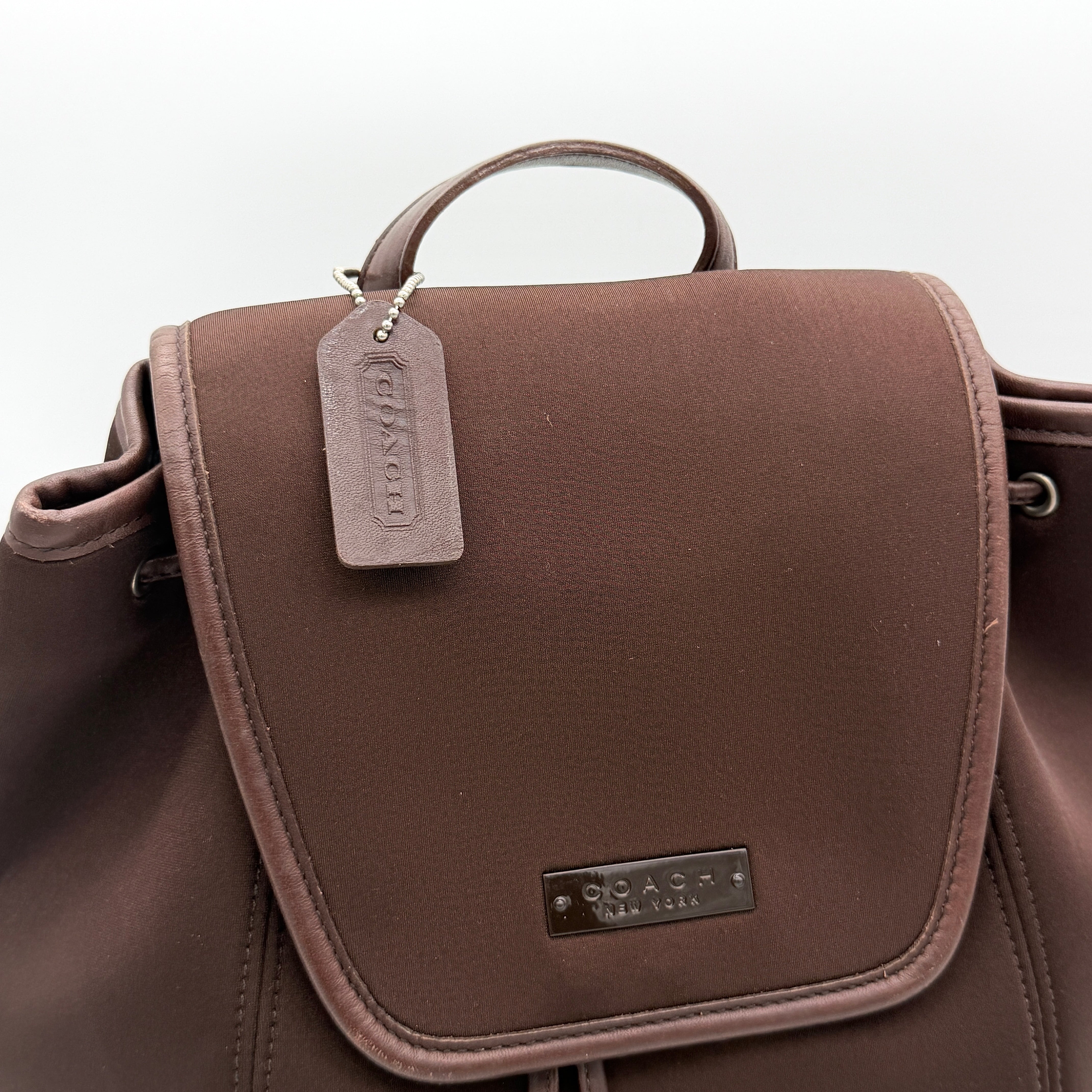 Coach brown neoprene drawstring backpack with leather trim, silver-tone hardware, and adjustable straps shown against a white background.