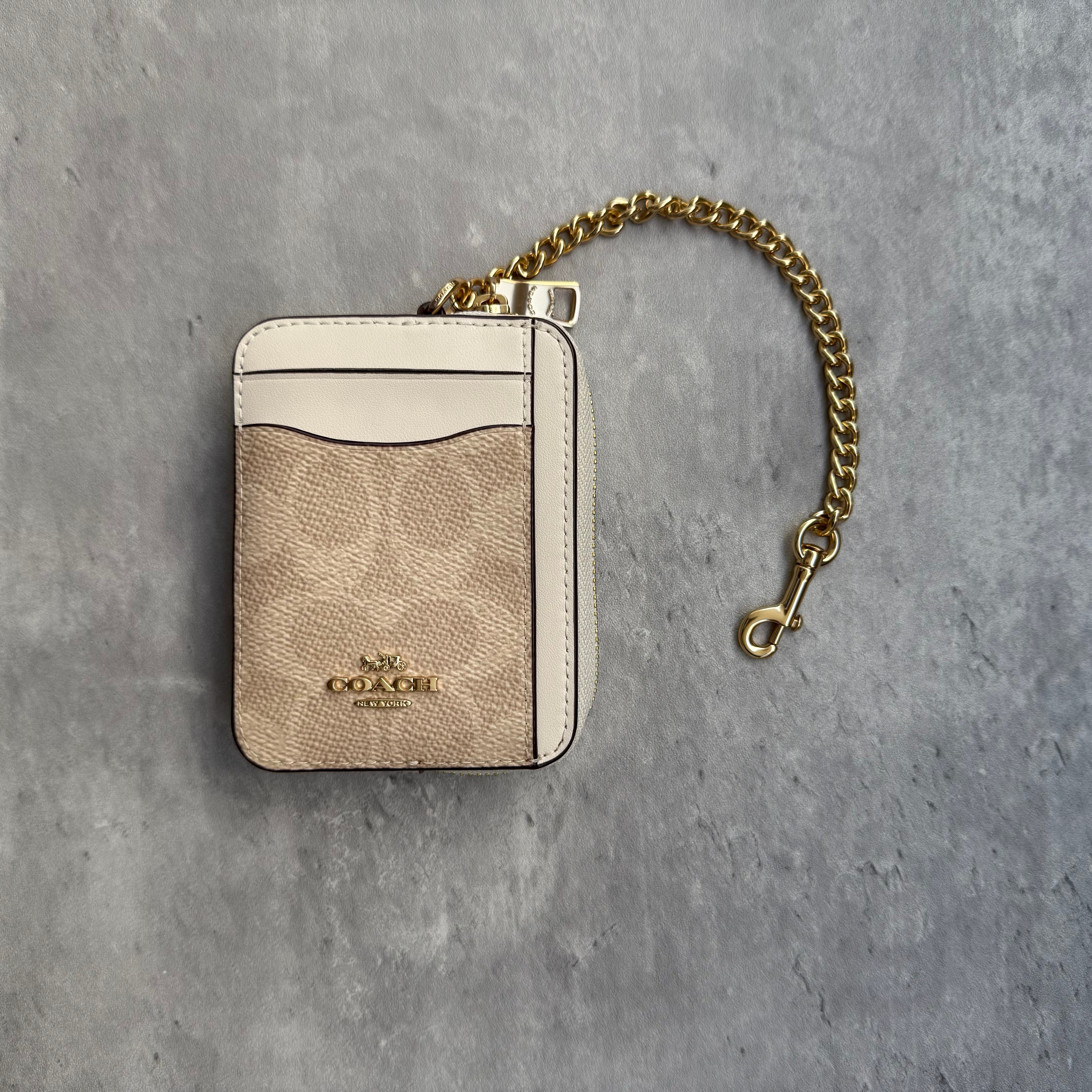 Coach Signature Chain Zip Cardholder in beige coated canvas with white leather trim, featuring gold-tone chain, zip closure, ID window, and Coach logo.
