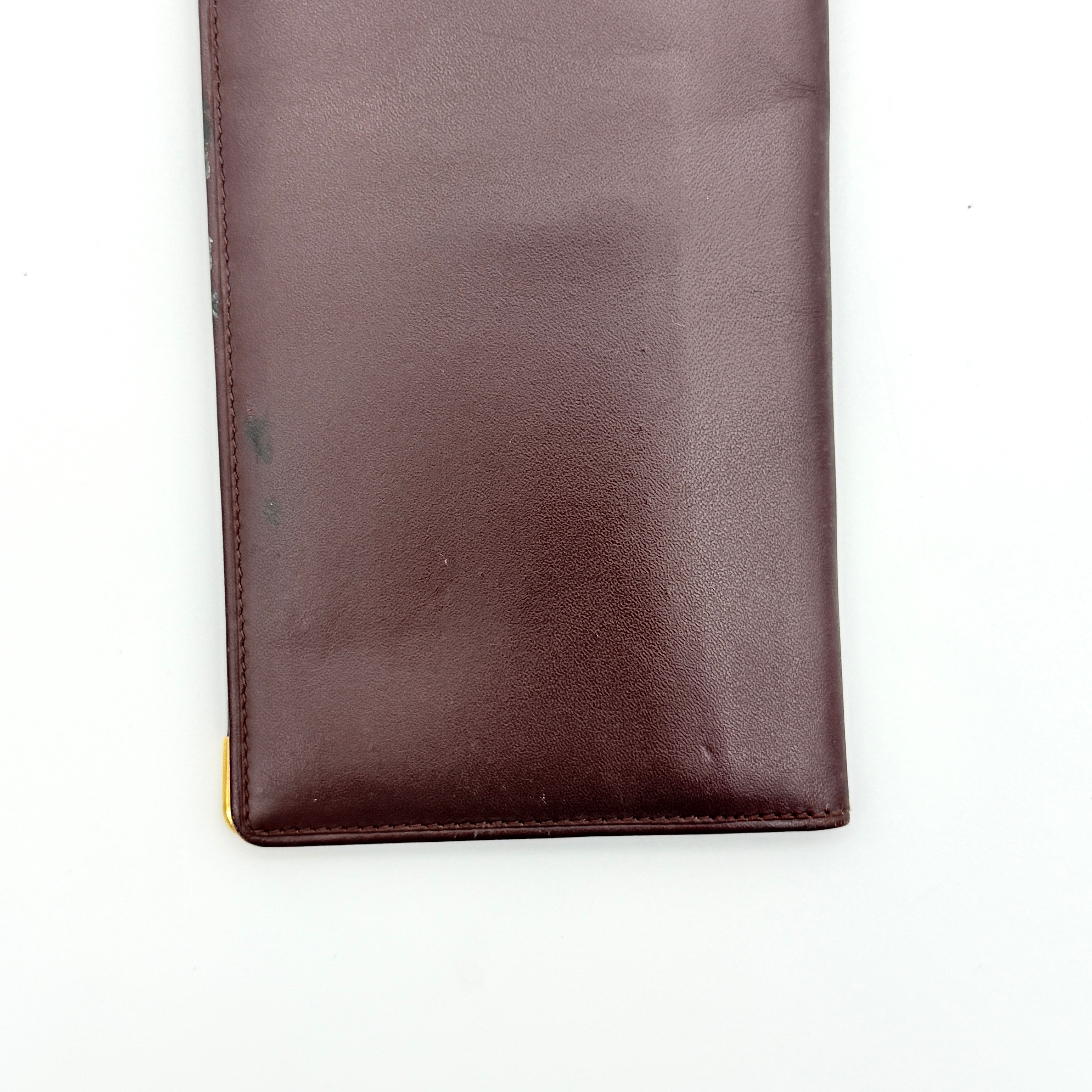 Cartier Must de Cartier bifold long wallet in burgundy leather with gold-tone corner accents, shown closed against a white background.