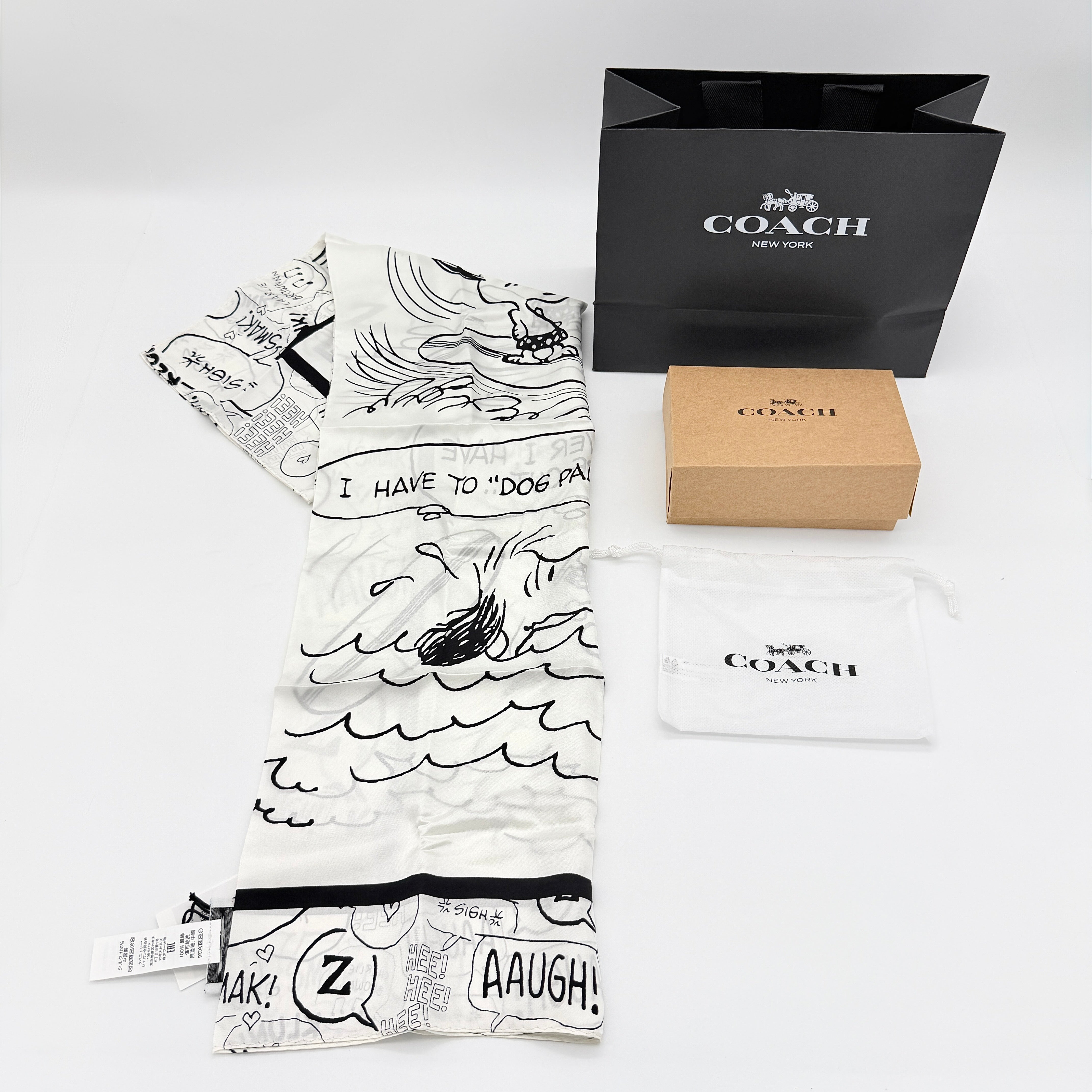 Coach x Peanuts black and white silk scarf featuring a Snoopy surfing comic print, shown with Coach packaging and tags.