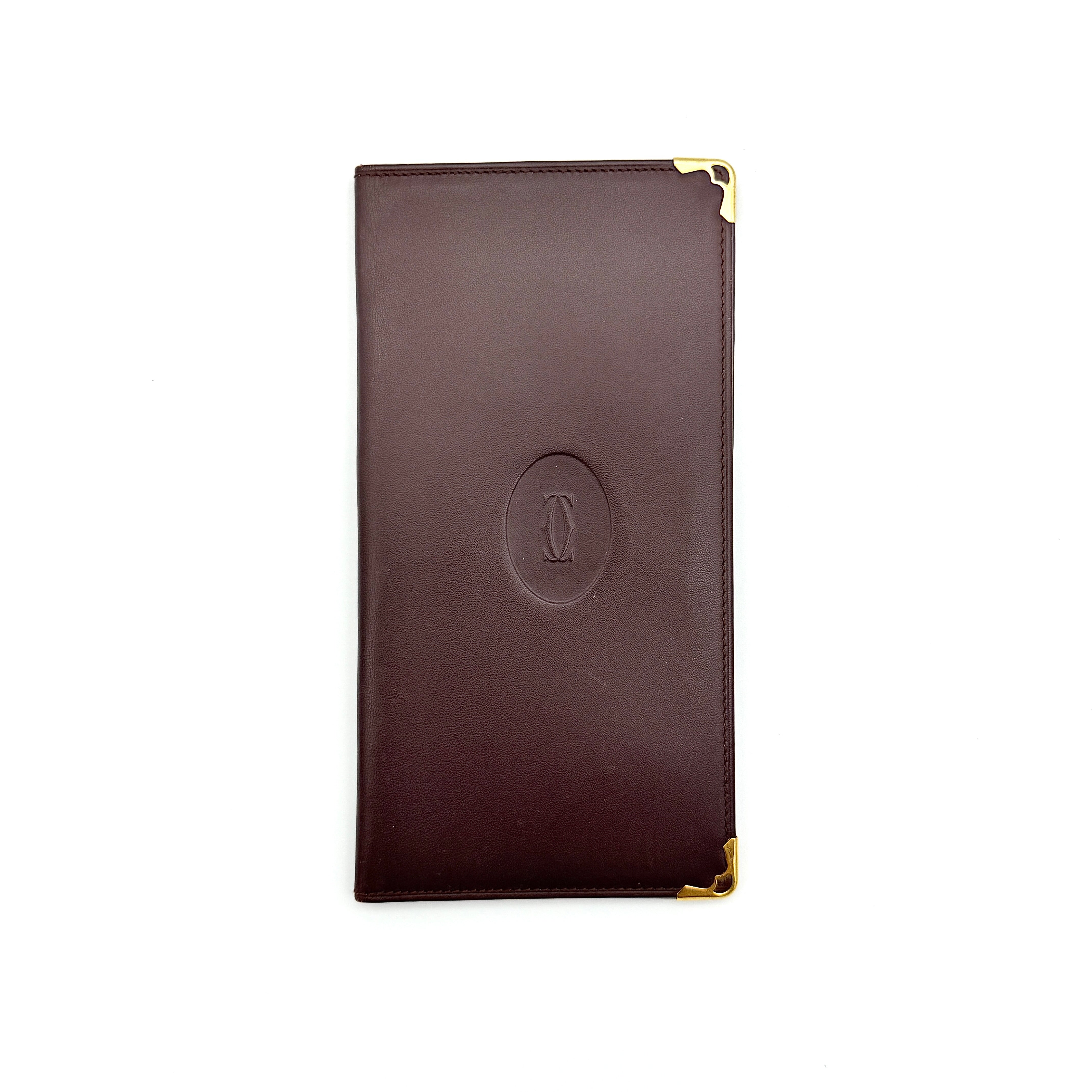 Cartier Must de Cartier bifold long wallet in burgundy leather with gold-tone corner accents, shown closed against a white background.