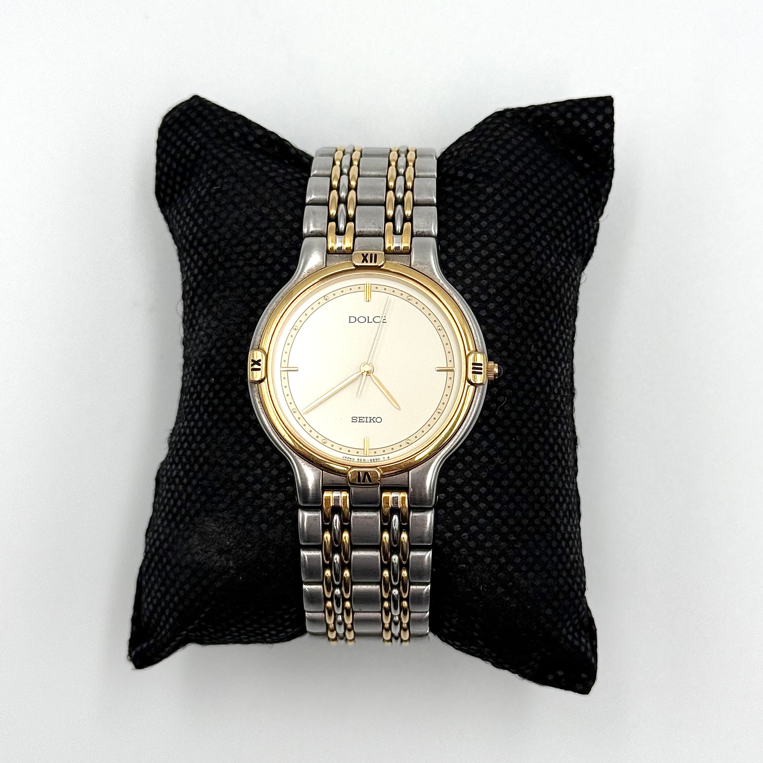 Seiko Dolce 5E31-6B90 watch in stainless steel with gold-tone accents and a cream dial, displayed on a black cushion.