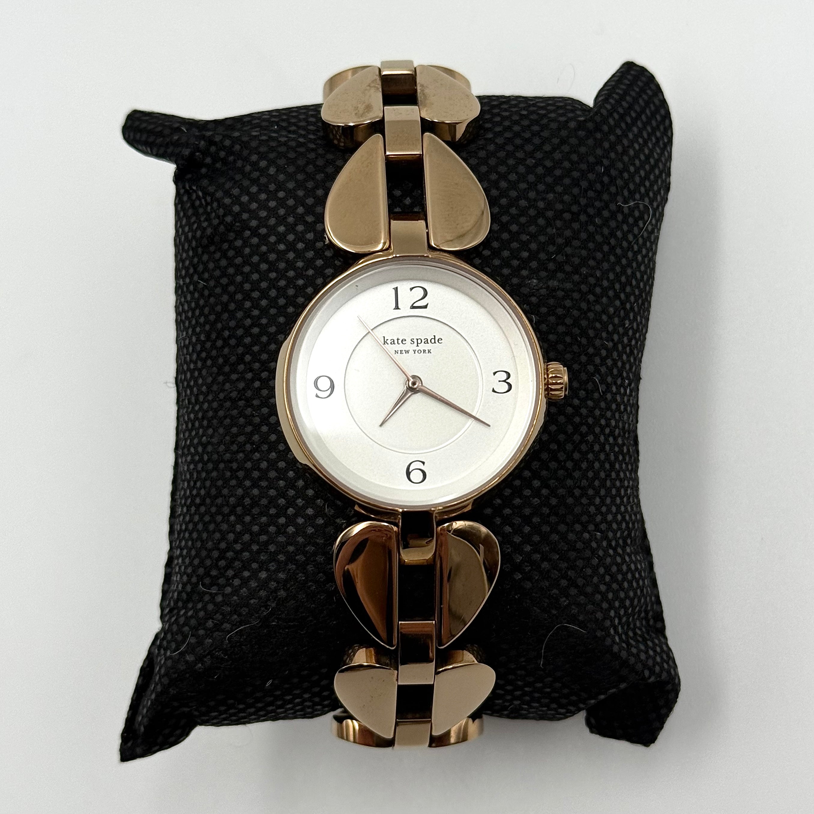 Kate Spade Annadale gold bracelet watch with white dial and heart-shaped links displayed on a black cushion.