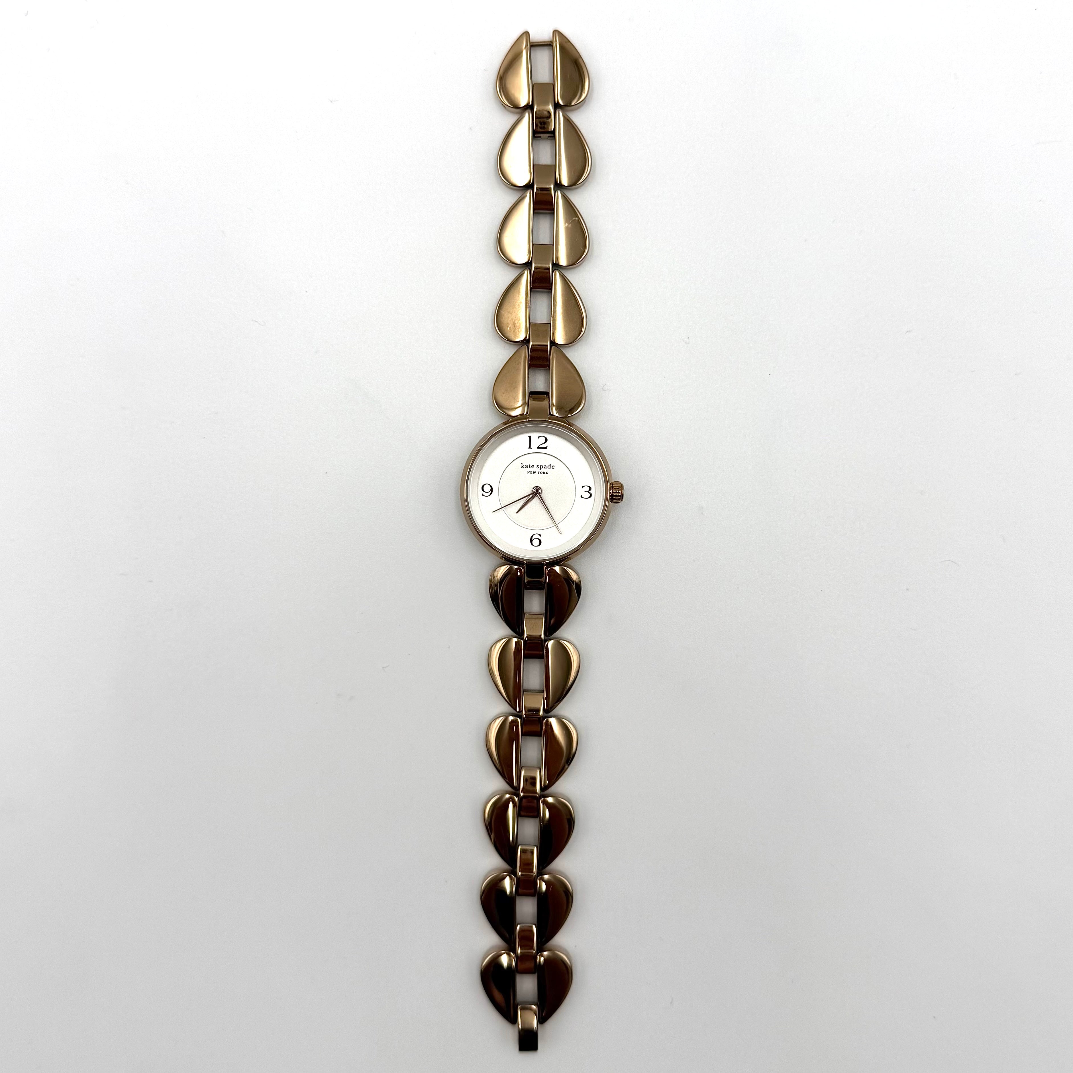 Kate Spade Annadale gold bracelet watch with white dial and heart-shaped links displayed on a black cushion.