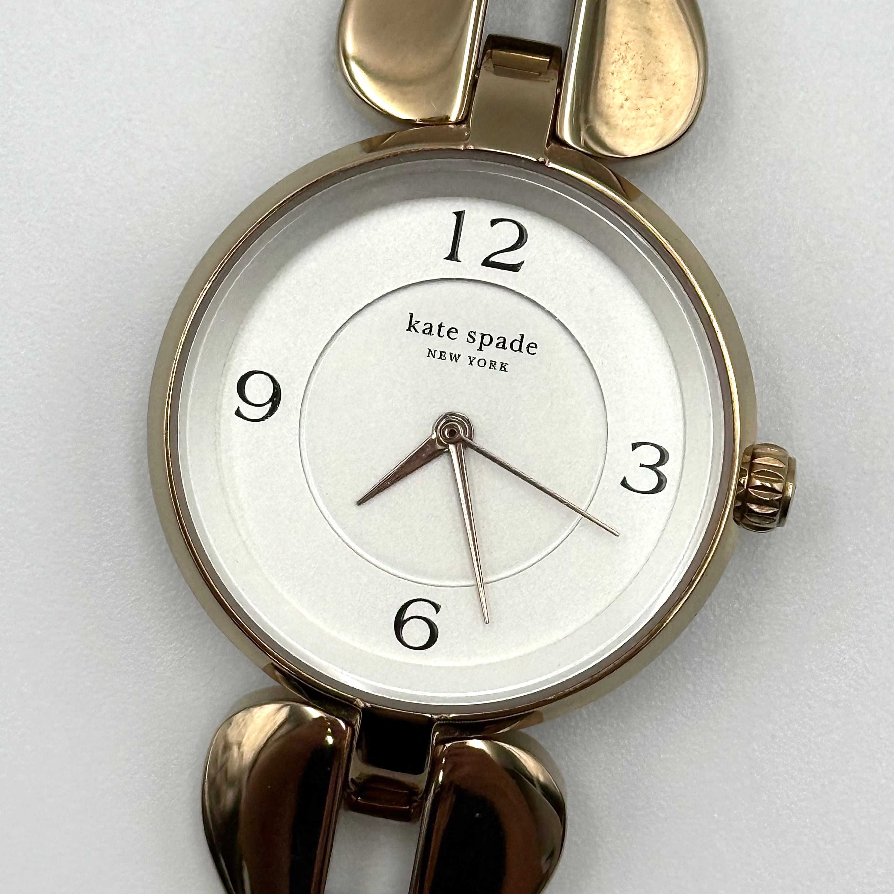 Kate Spade Annadale gold bracelet watch with white dial and heart-shaped links displayed on a black cushion.