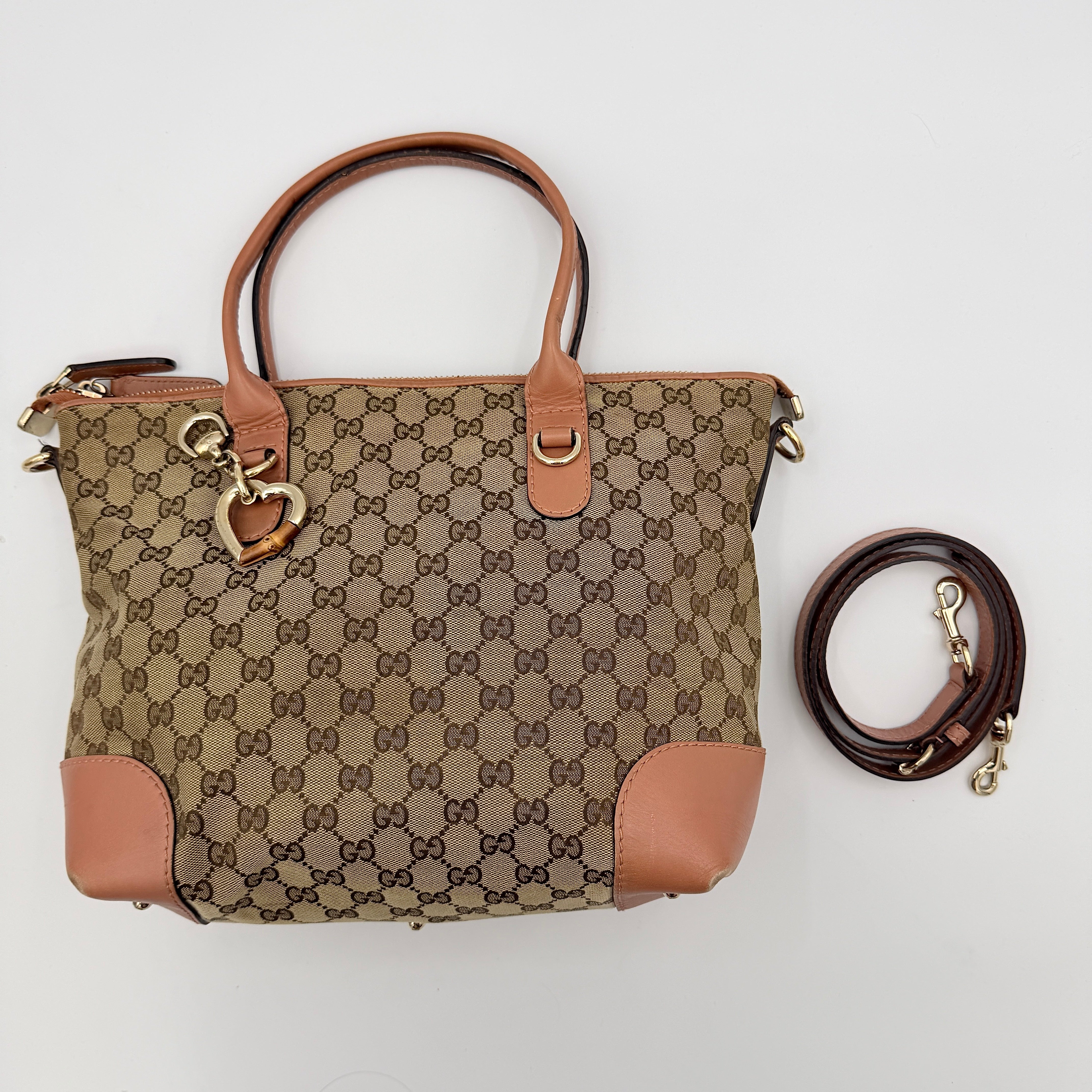 Gucci beige GG monogram shoulder bag with orange leather trim, heart bit charm, and detachable strap displayed on a white background.