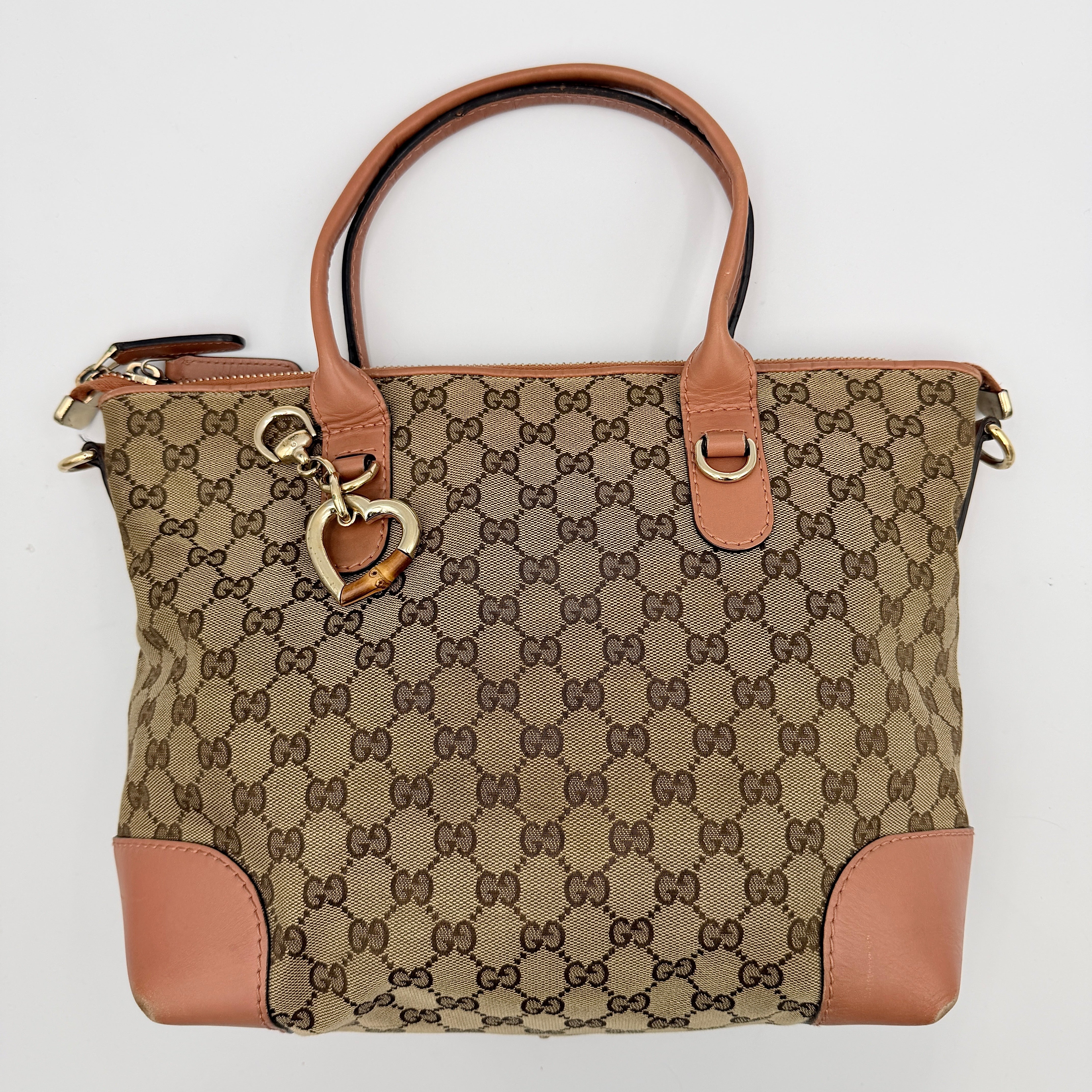 Gucci beige GG monogram shoulder bag with orange leather trim, heart bit charm, and detachable strap displayed on a white background.