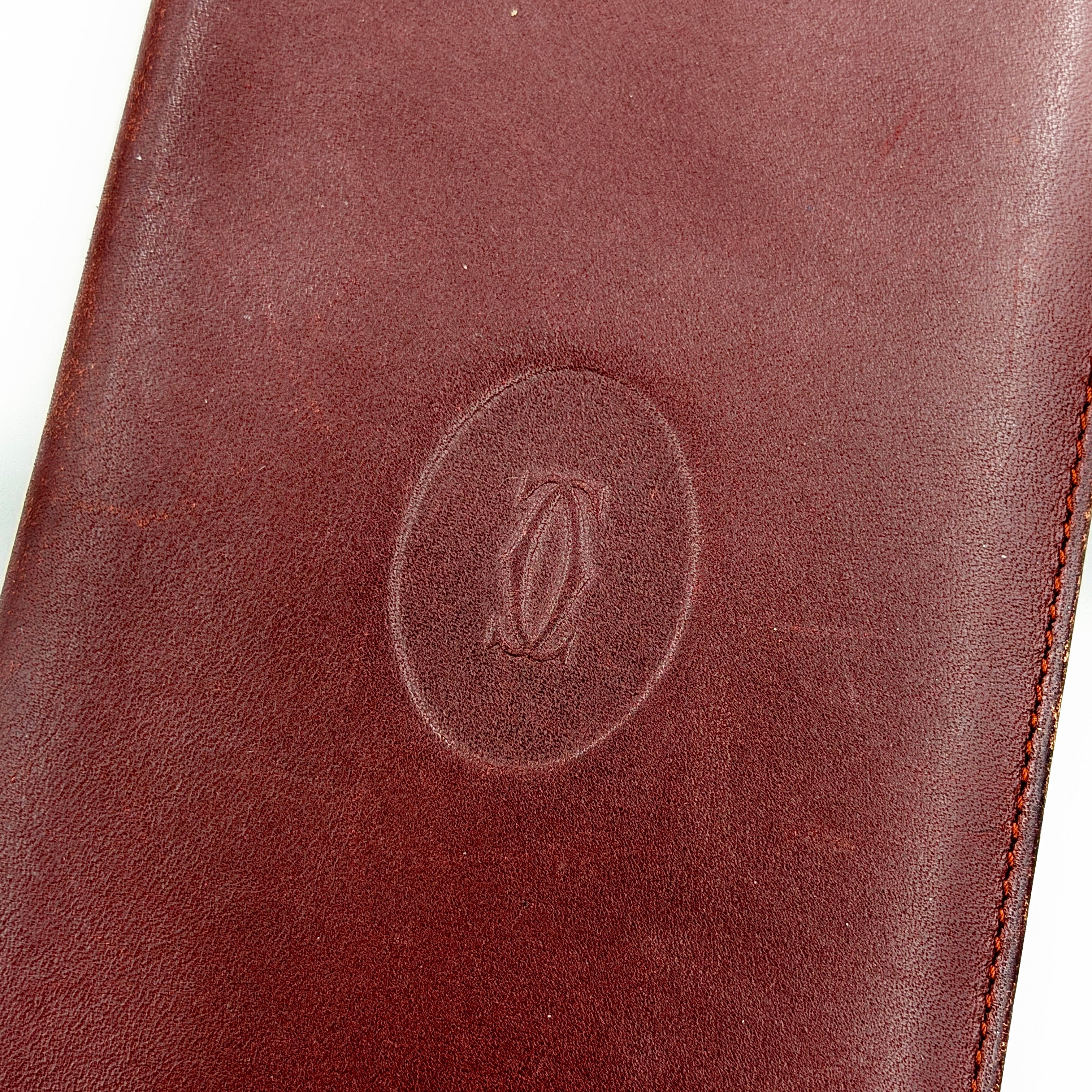 The Cartier Must de Cartier long bifold wallet in rich bordeaux calfskin with gold-tone corner accents and an embossed logo.