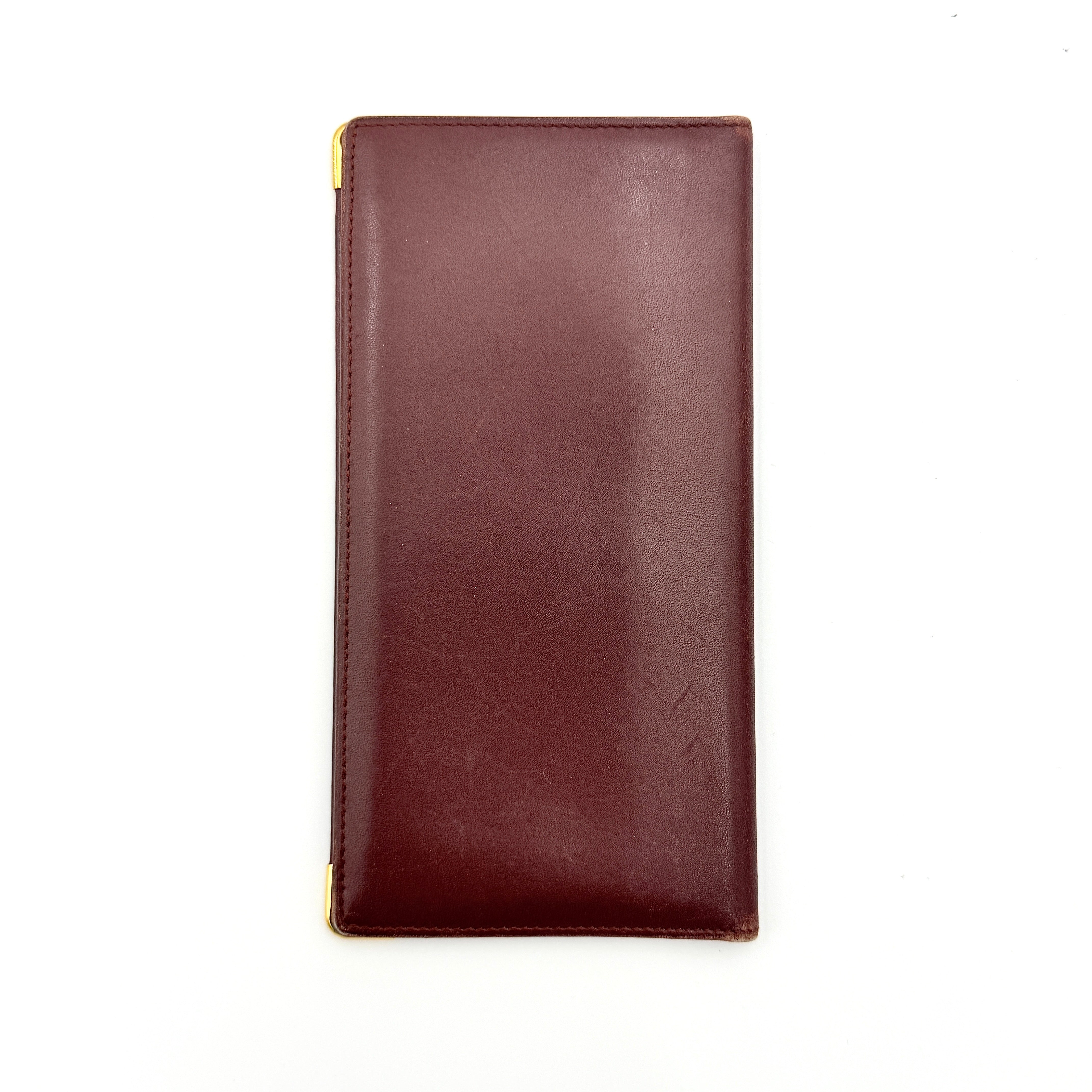 The Cartier Must de Cartier long bifold wallet in rich bordeaux calfskin with gold-tone corner accents and an embossed logo.