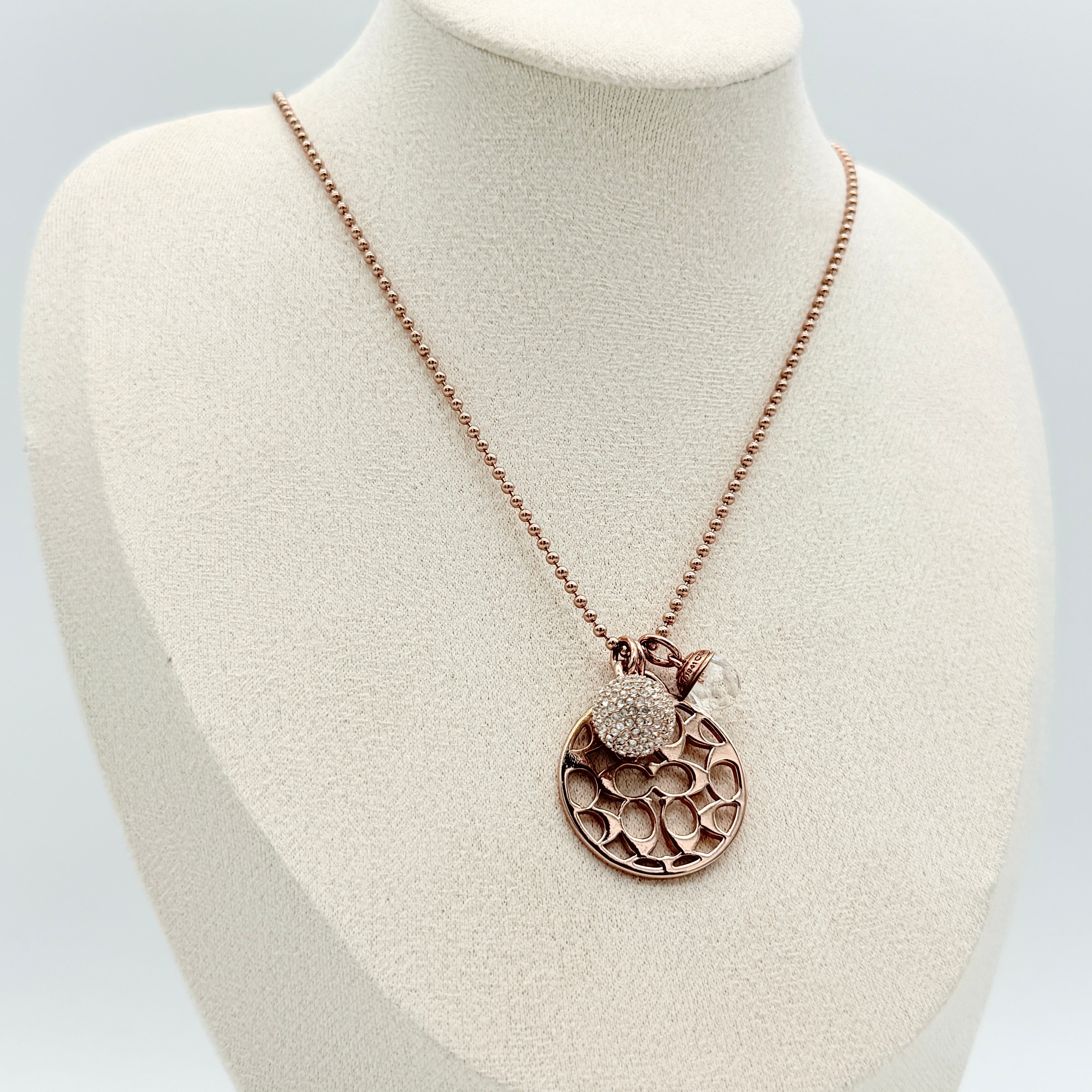 Coach rose gold necklace with signature cut-out medallion, crystal pavé ball and faceted charm on a ball chain.