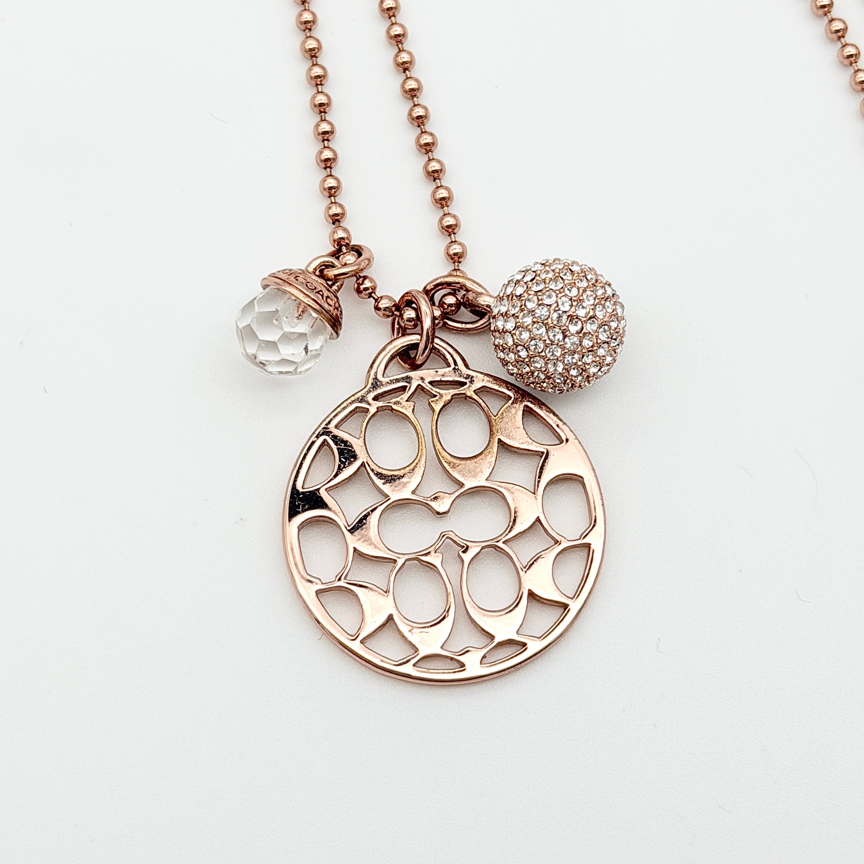 Coach rose gold necklace with signature cut-out medallion, crystal pavé ball and faceted charm on a ball chain.