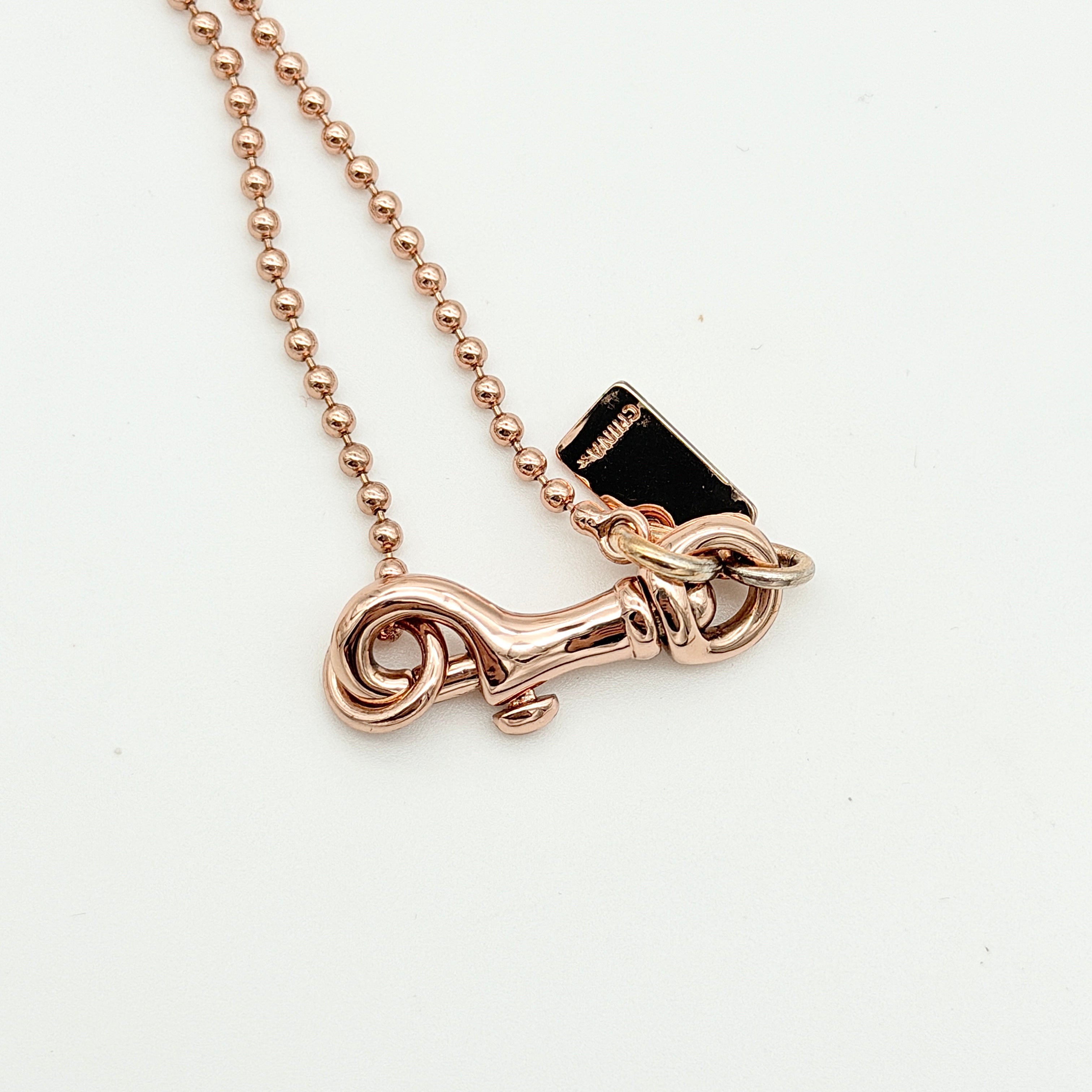 Coach rose gold necklace with signature cut-out medallion, crystal pavé ball and faceted charm on a ball chain.