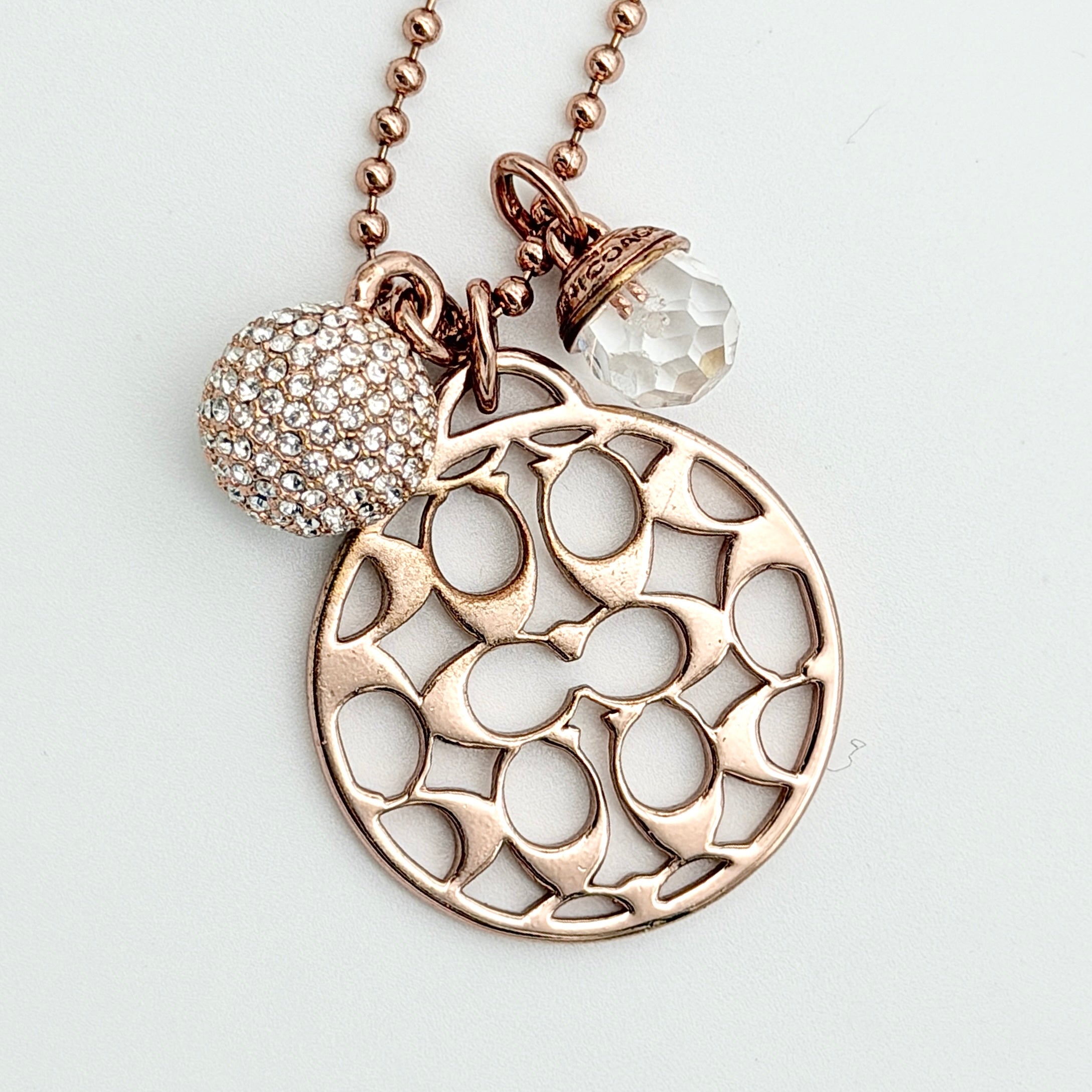 Coach rose gold necklace with signature cut-out medallion, crystal pavé ball and faceted charm on a ball chain.