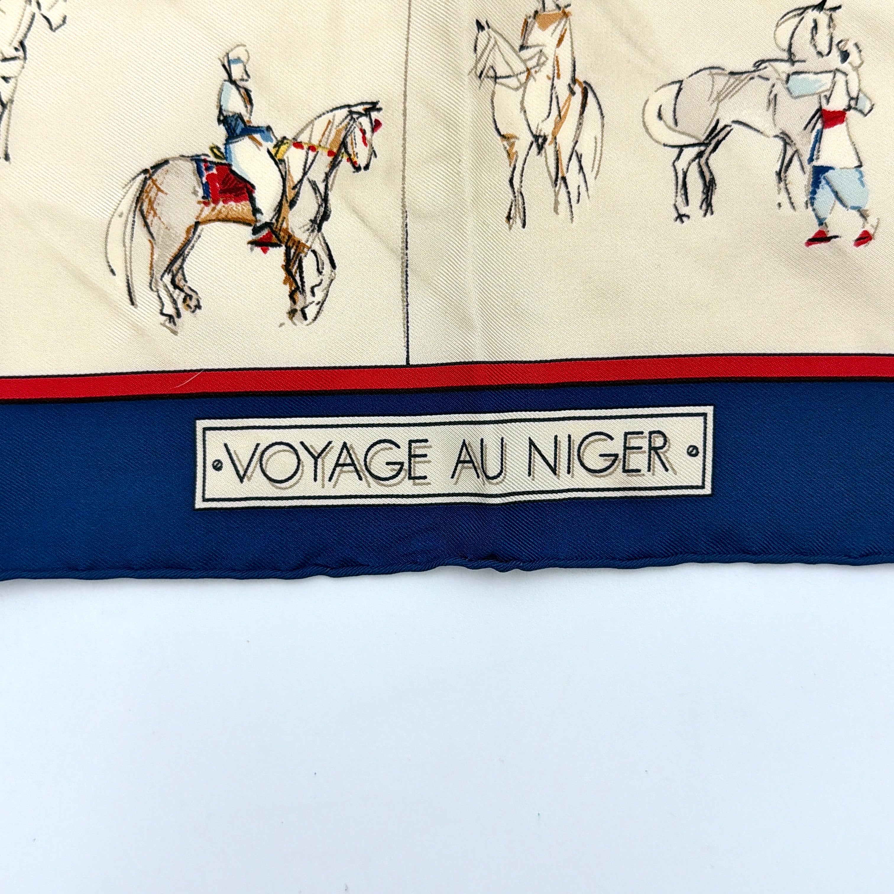 Hermès Voyage au Niger silk scarf in navy blue with cream center, featuring illustrative equestrian sketches bordered in red.