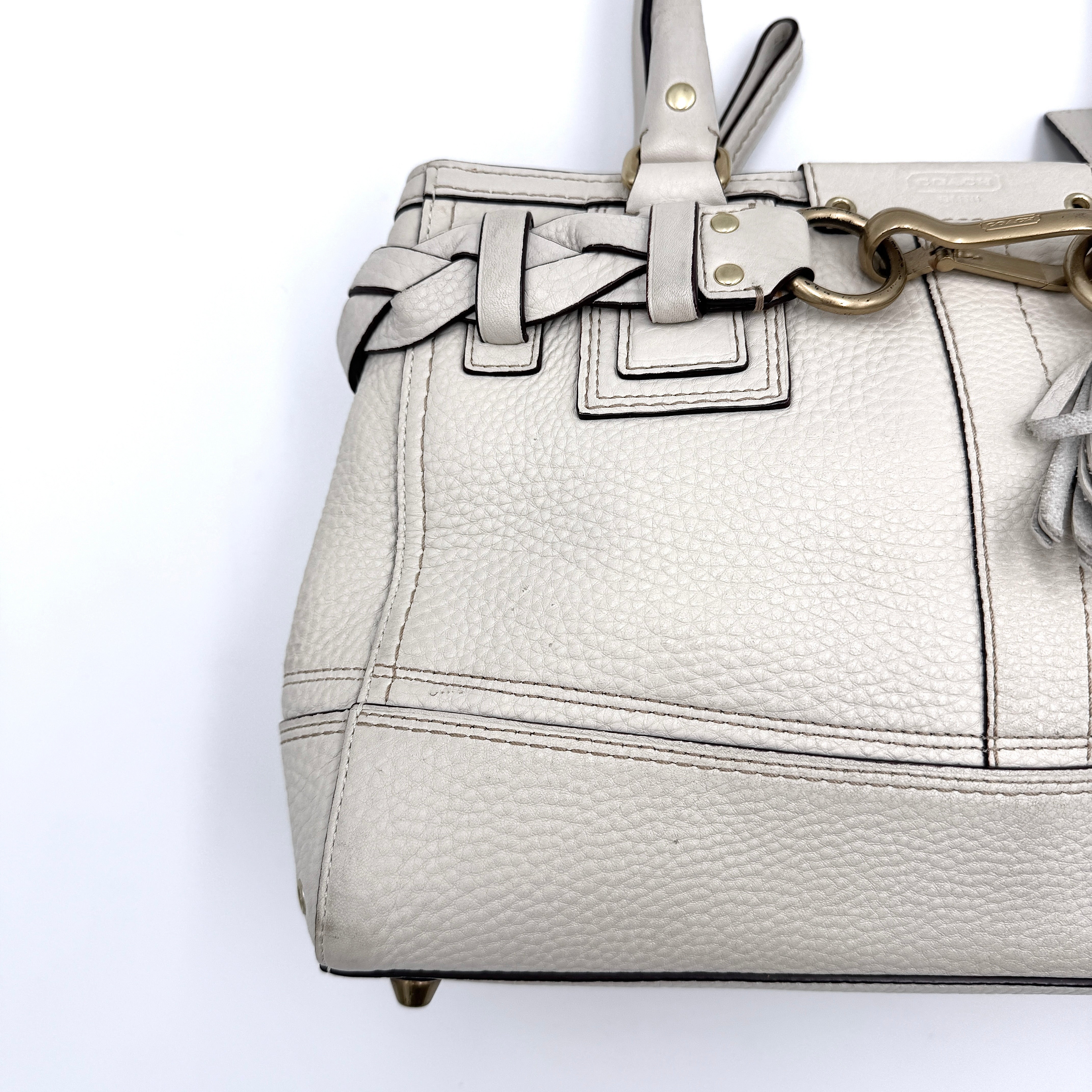 Coach Hampton Carryall in ivory leather with braided trim, brass dogleash clip, tassel, and top handles displayed against a white background.