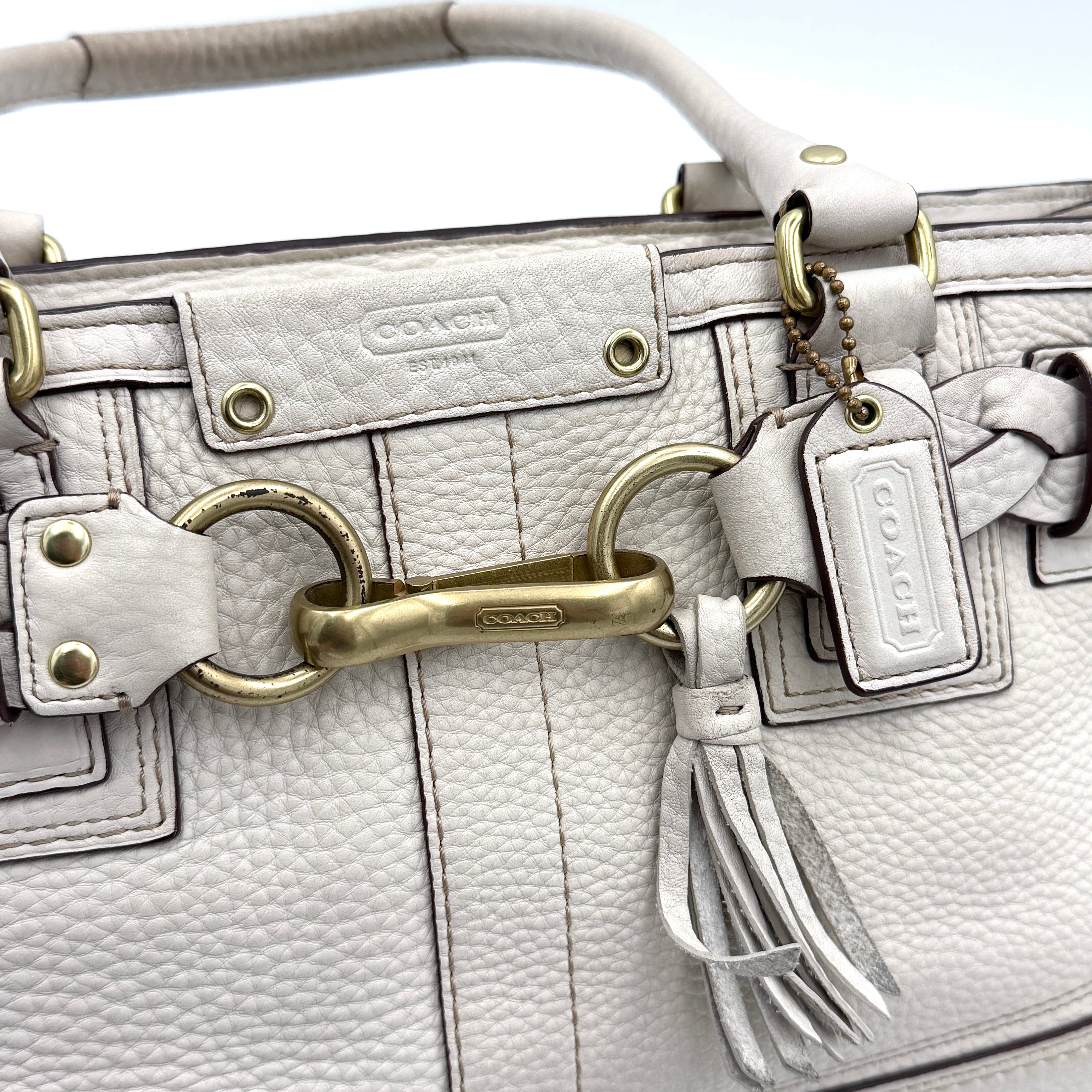 Coach Hampton Carryall in ivory leather with braided trim, brass dogleash clip, tassel, and top handles displayed against a white background.