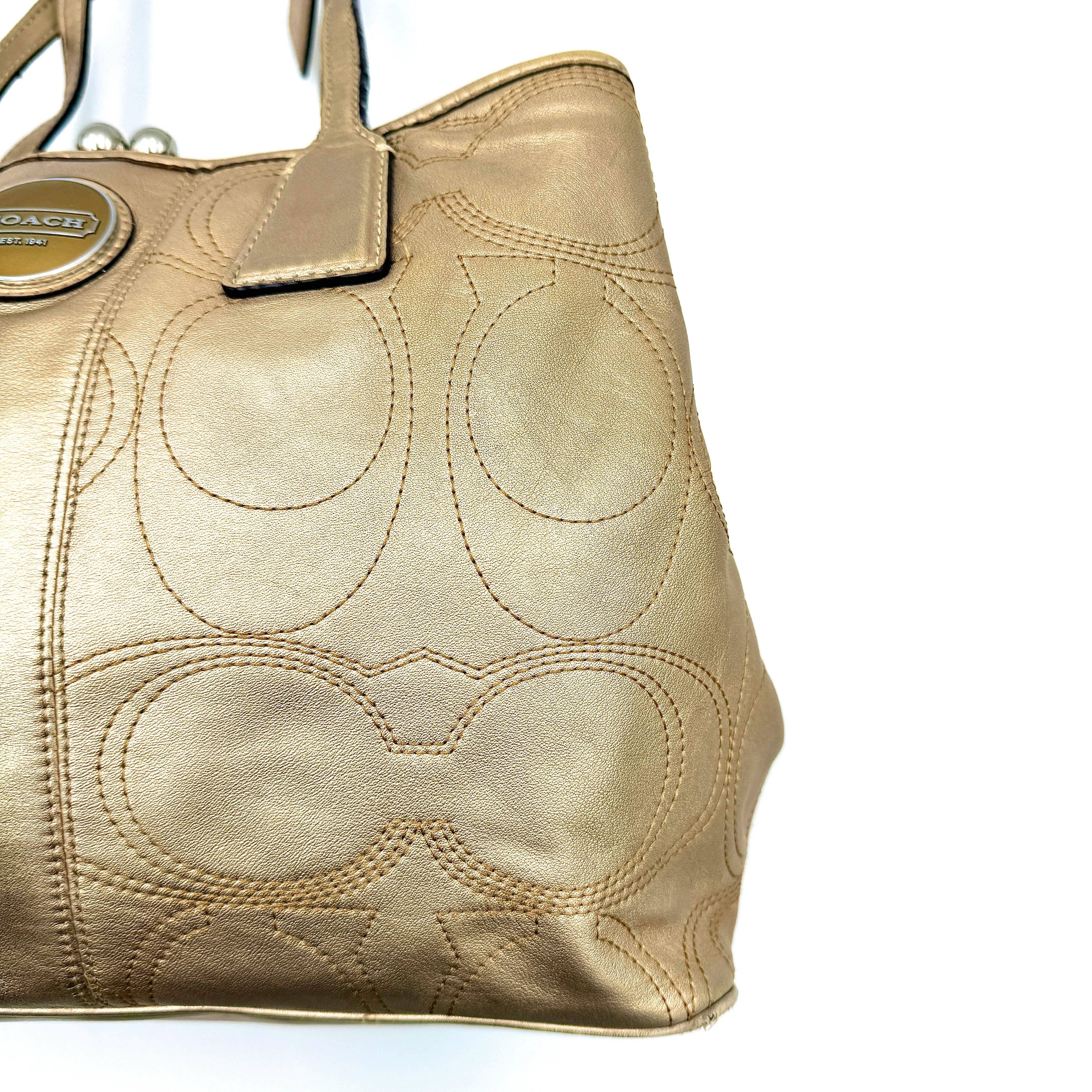 Coach metallic gold shoulder bag with signature embossed leather, double top handles, and a kisslock closure set against a clean background.