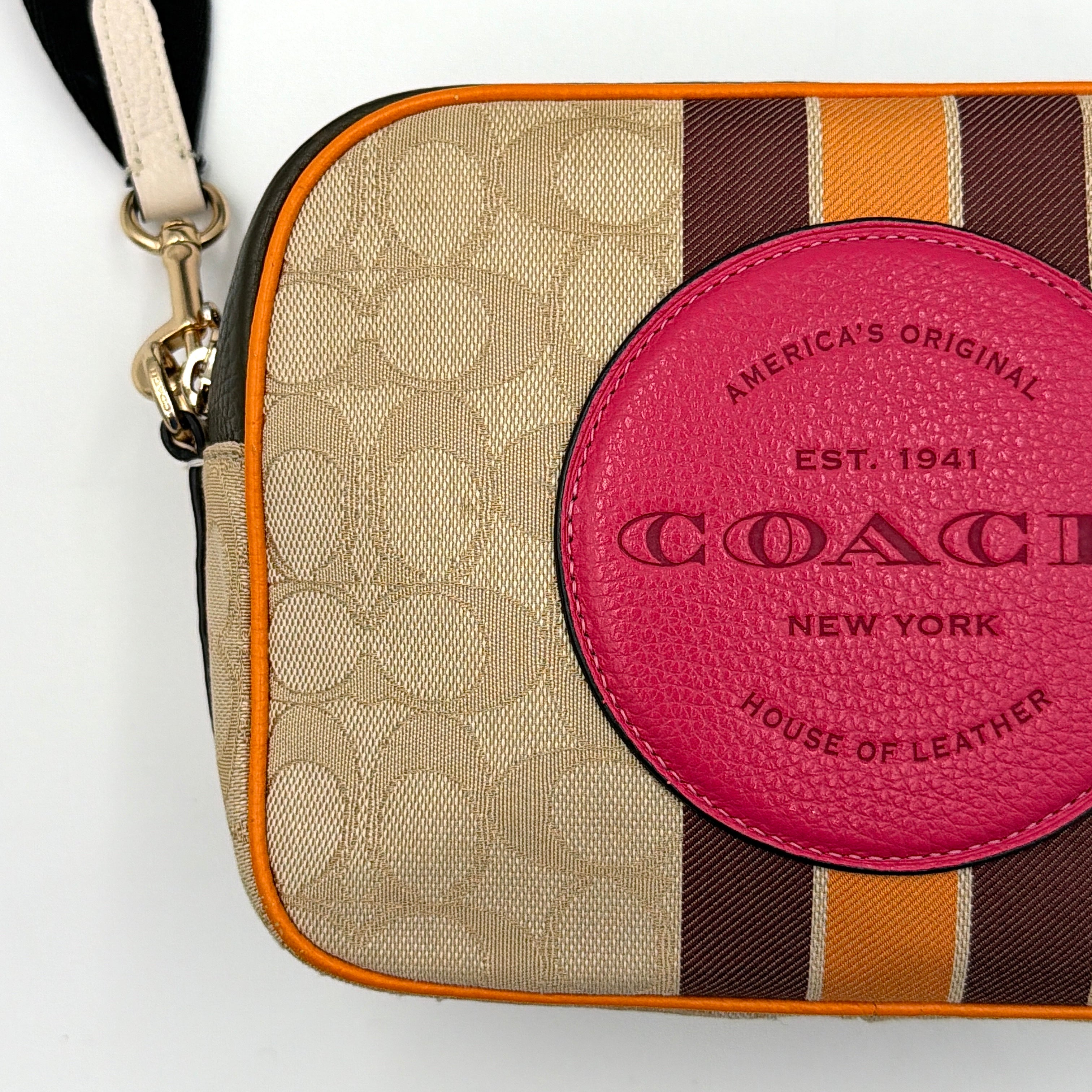 Coach Signature Dempsey Camera Bag in monogram canvas with bold pink logo patch, orange stripes, and wide black strap.
