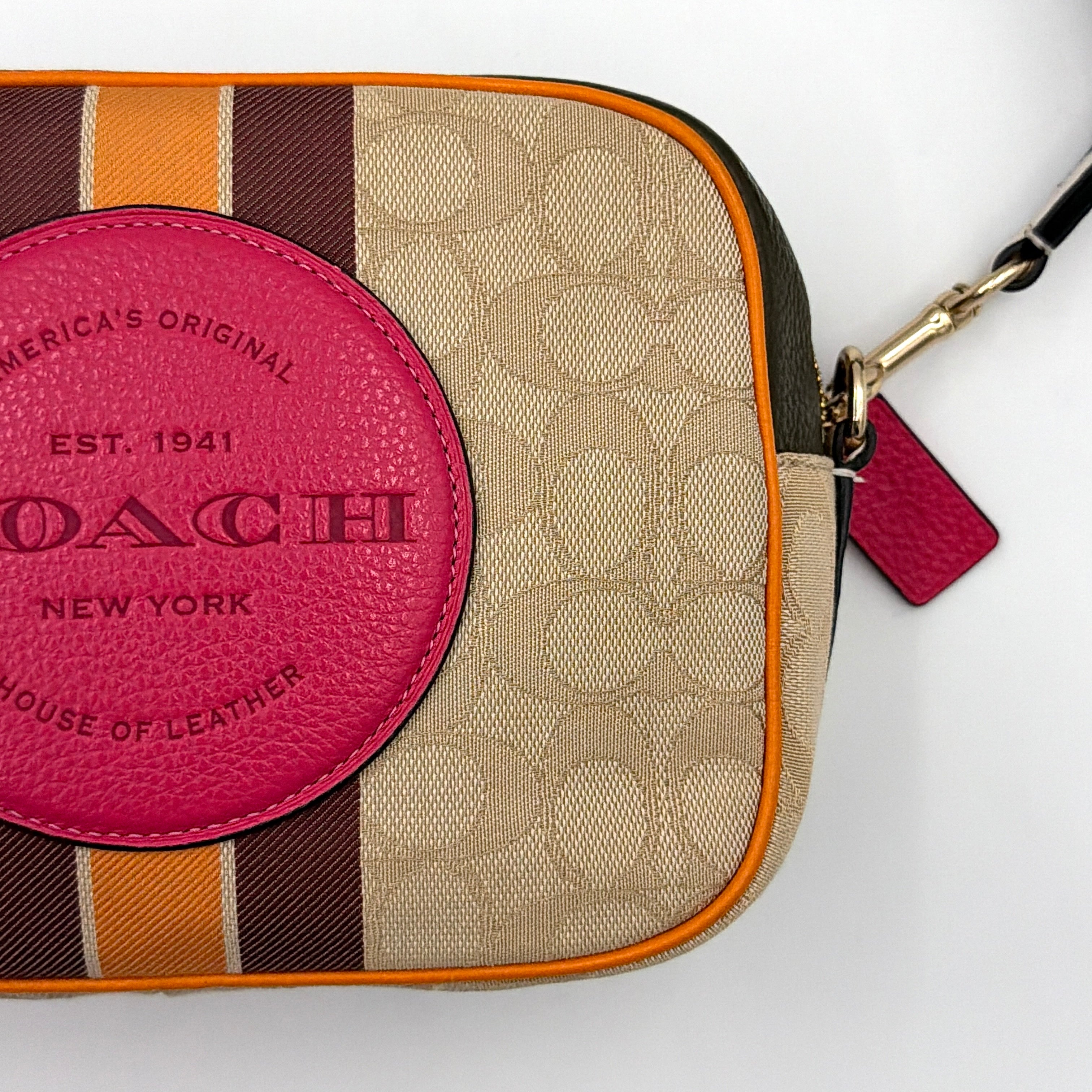 Coach Signature Dempsey Camera Bag in monogram canvas with bold pink logo patch, orange stripes, and wide black strap.