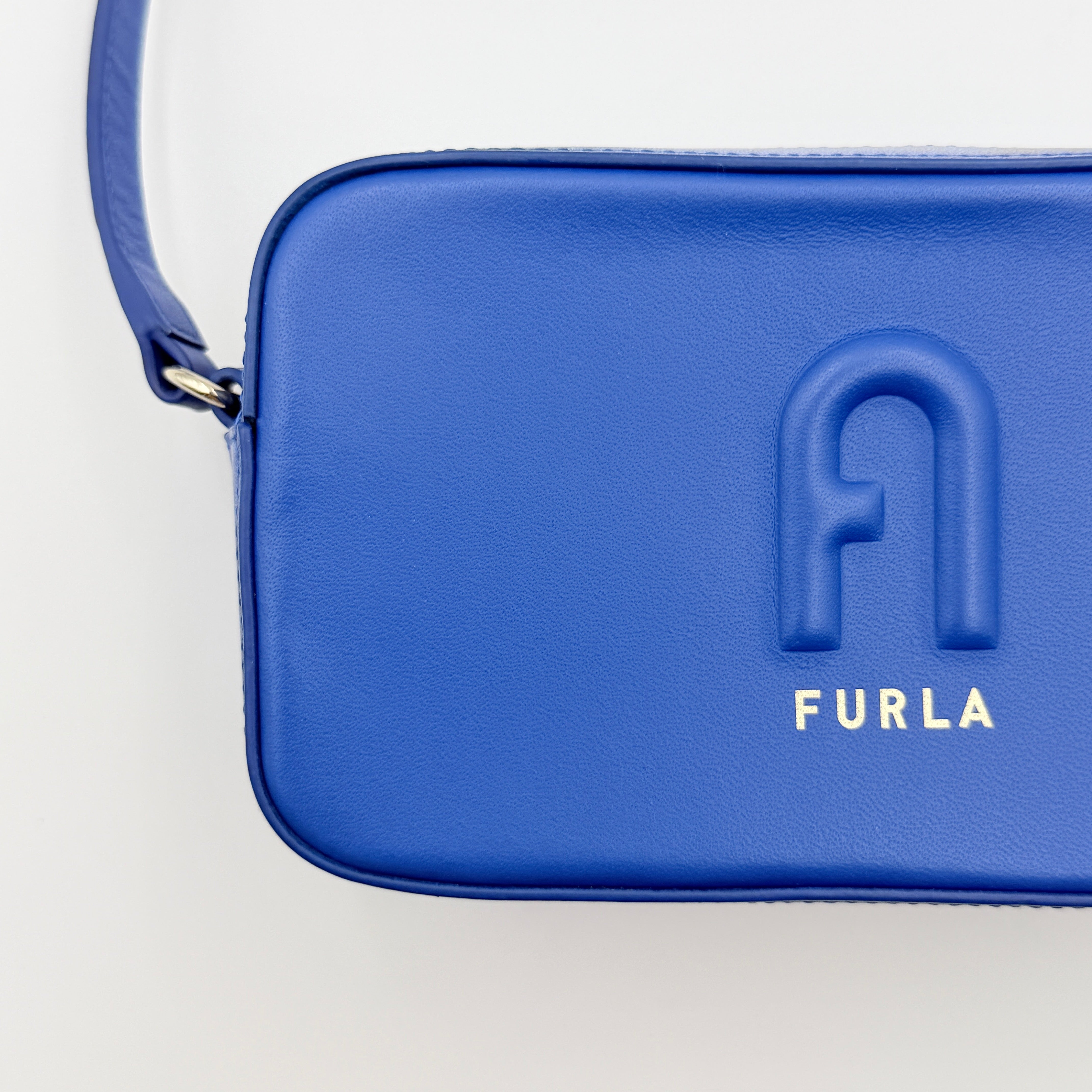 Furla Primula Mini Camera Crossbody Bag in vibrant blue leather with embossed logo, shown with adjustable strap.