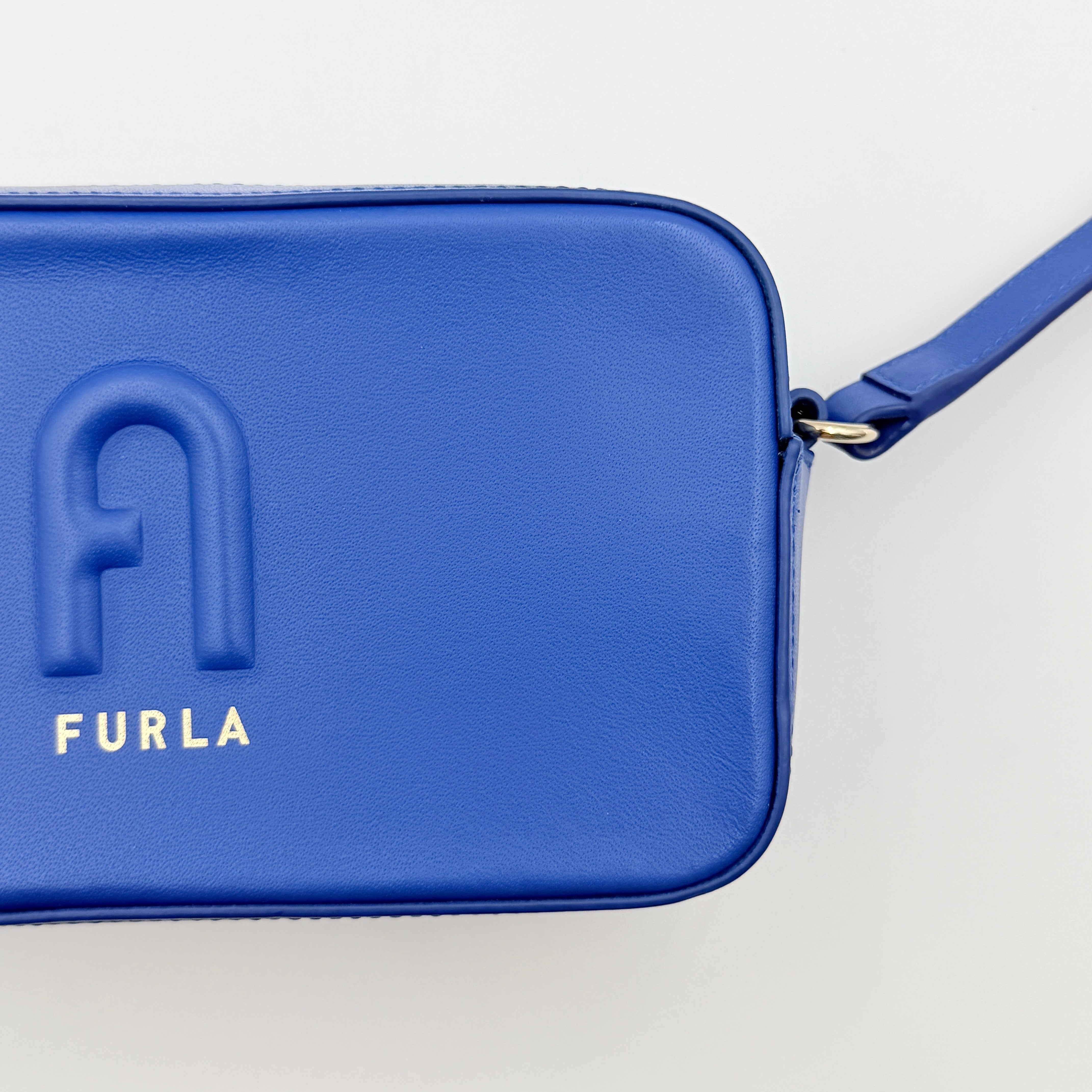 Furla Primula Mini Camera Crossbody Bag in vibrant blue leather with embossed logo, shown with adjustable strap.