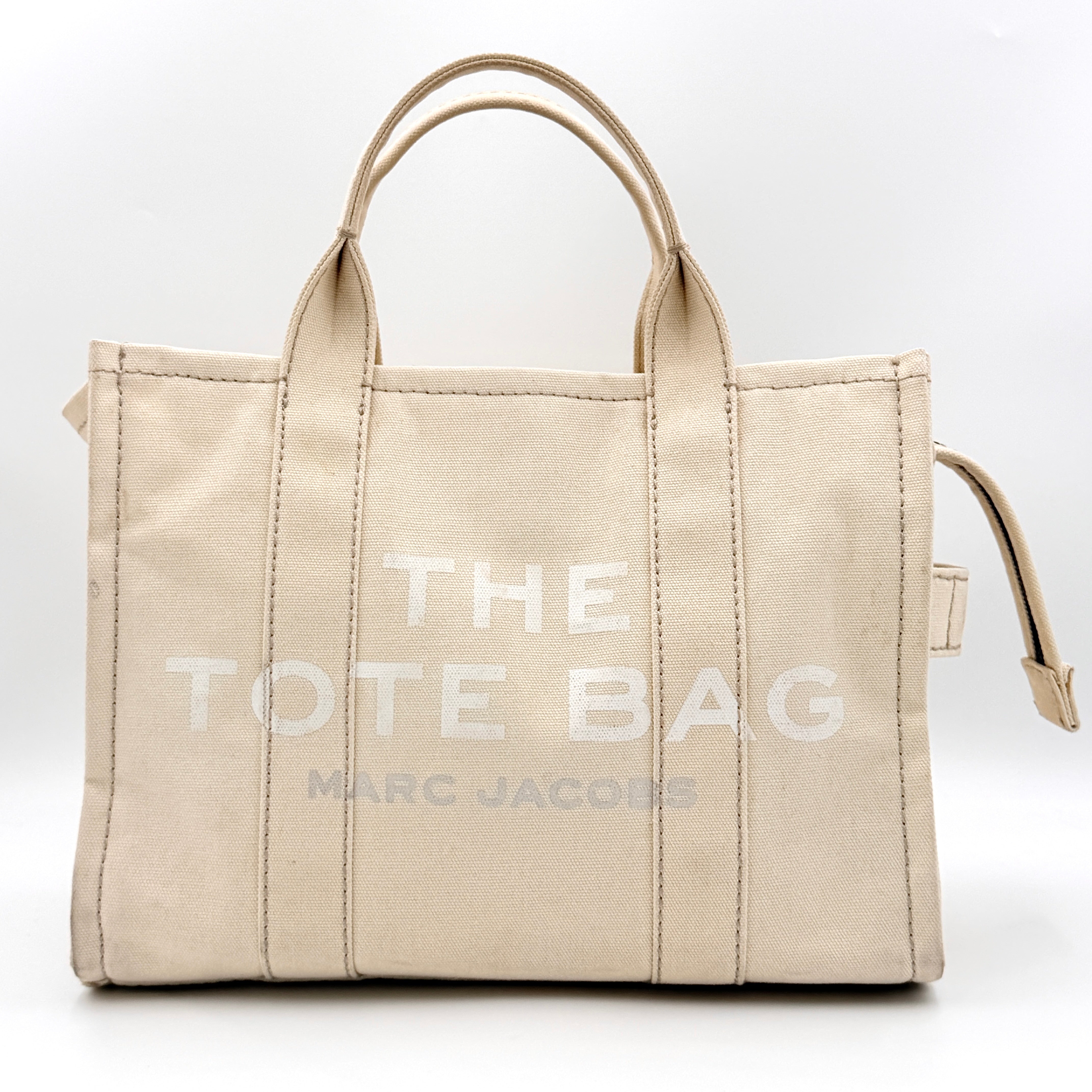 Marc Jacobs The Canvas Tote Bag in beige canvas with bold text, featuring top handles and zip closure.