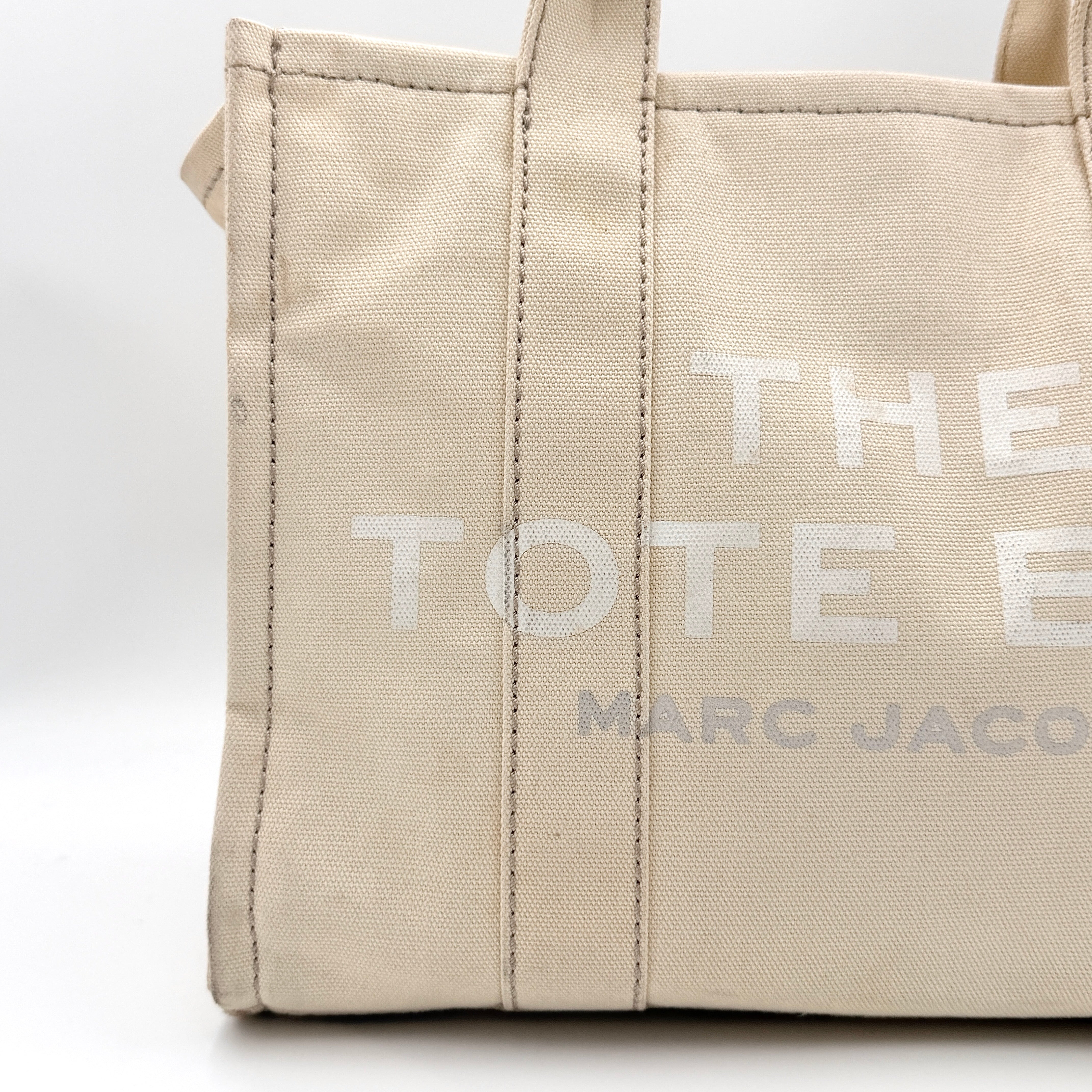 Marc Jacobs The Canvas Tote Bag in beige canvas with bold text, featuring top handles and zip closure.
