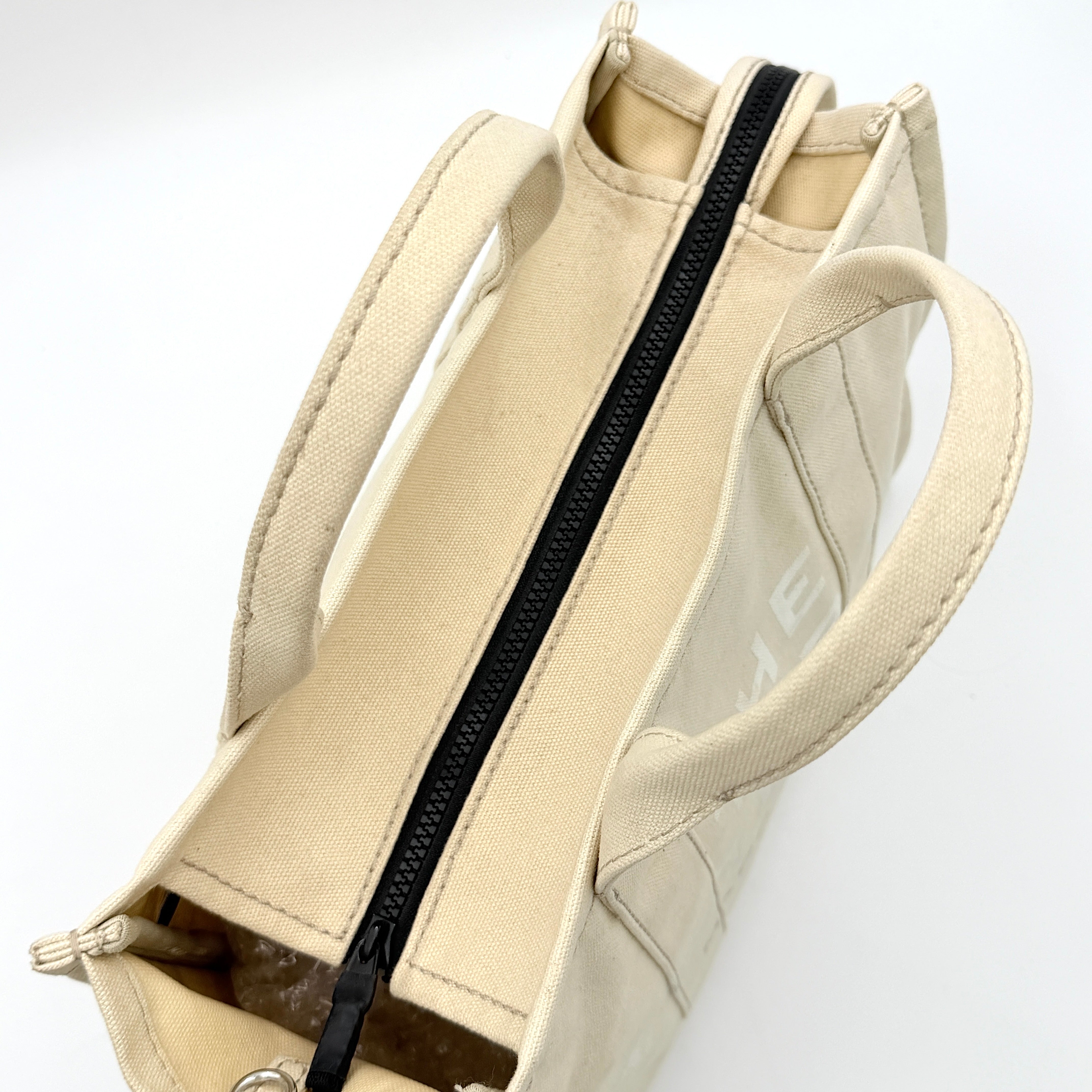Marc Jacobs The Canvas Tote Bag in beige canvas with bold text, featuring top handles and zip closure.