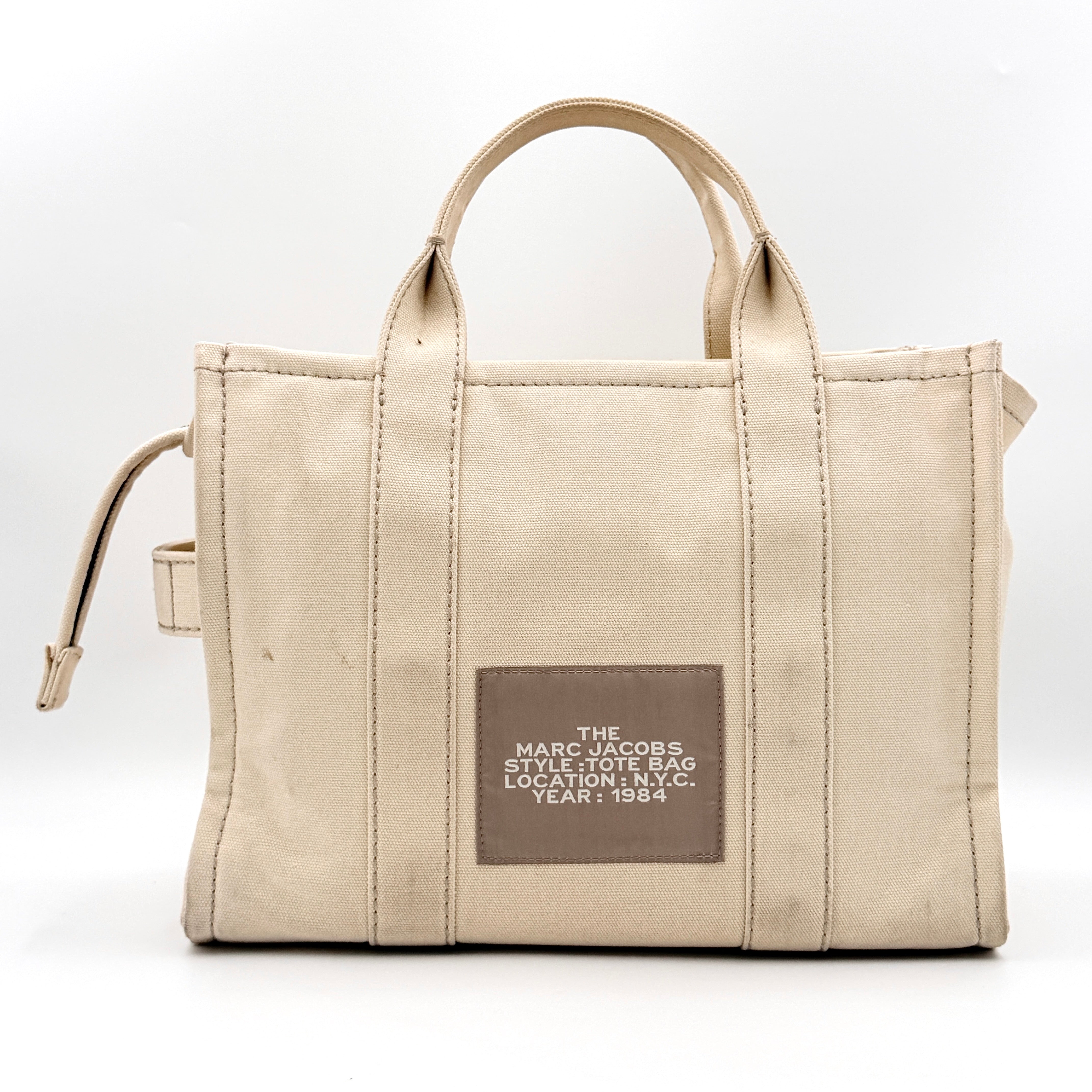 Marc Jacobs The Canvas Tote Bag in beige canvas with bold text, featuring top handles and zip closure.