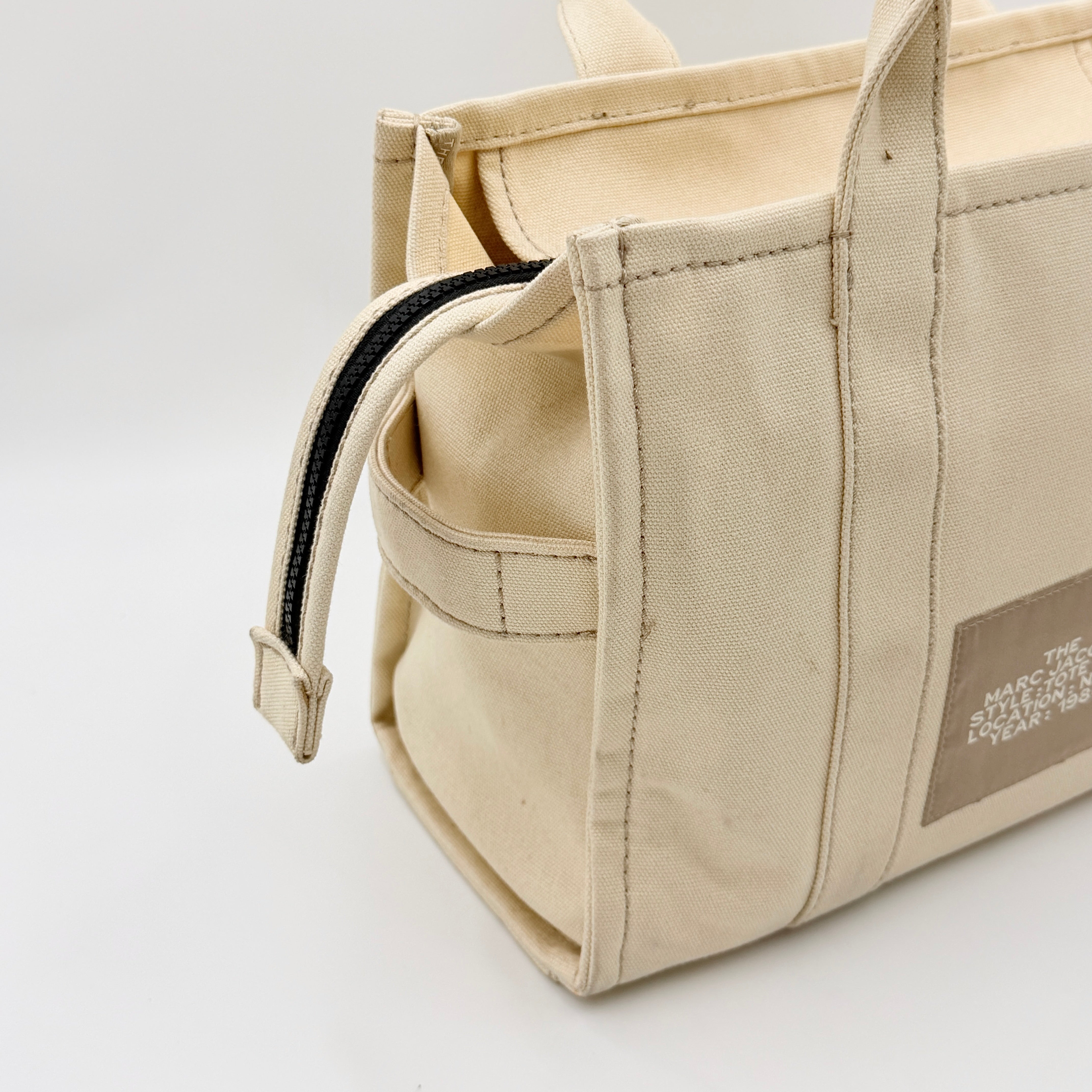 Marc Jacobs The Canvas Tote Bag in beige canvas with bold text, featuring top handles and zip closure.