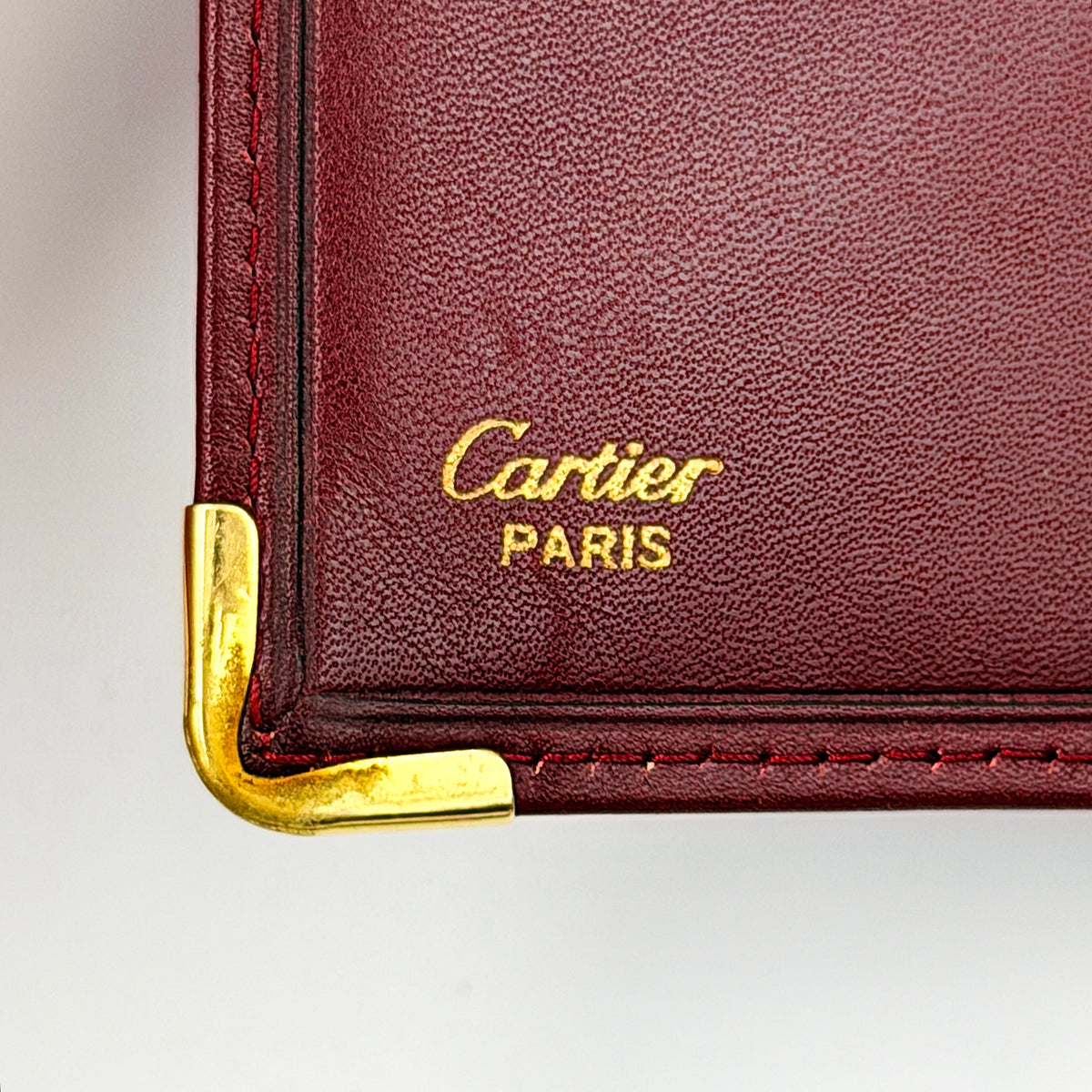 Cartier Must de Cartier bifold wallet in burgundy leather with gold-tone corner hardware and embossed logo, shown with authenticity card.