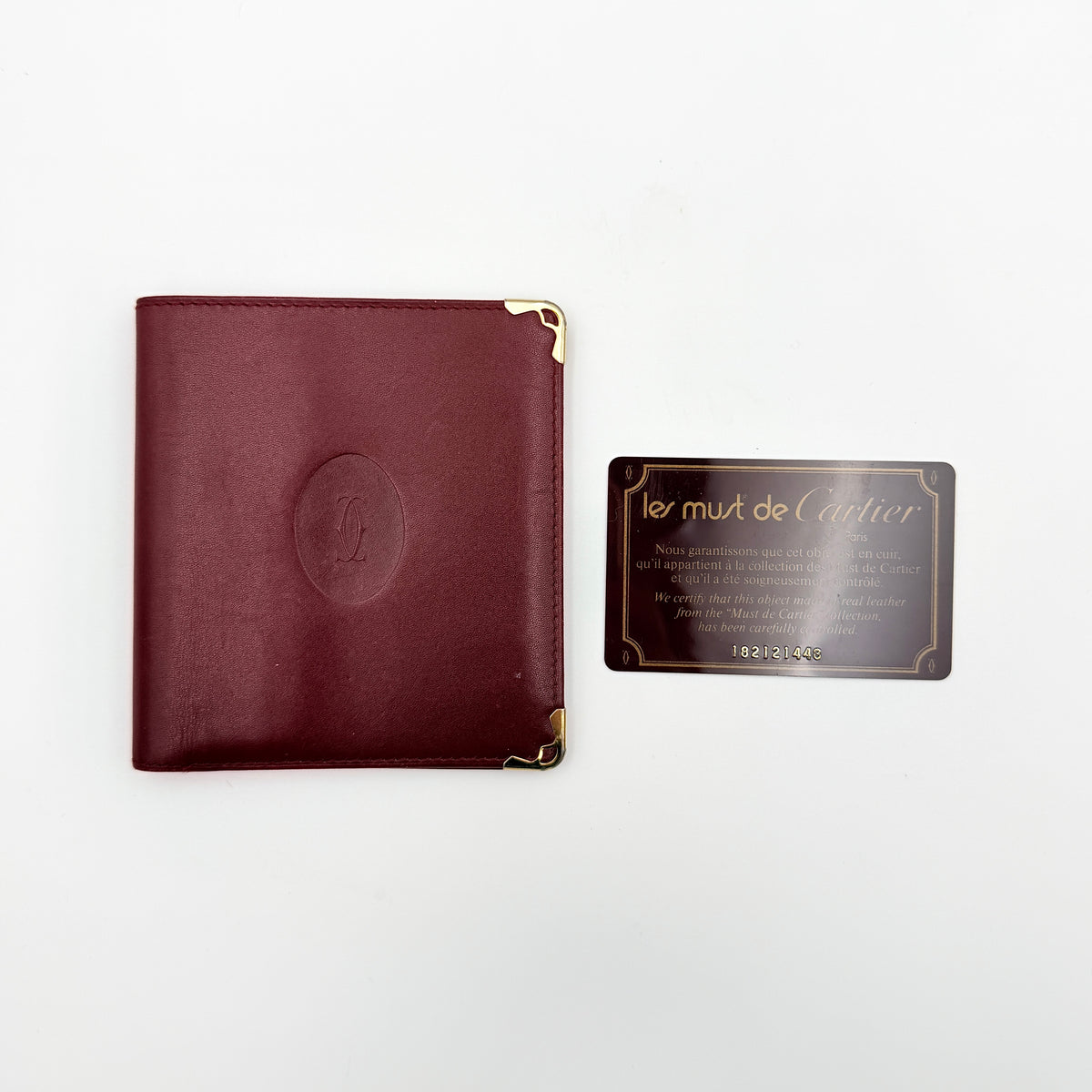 Cartier Must de Cartier bifold wallet in burgundy leather with gold-tone corner hardware and embossed logo, shown with authenticity card.