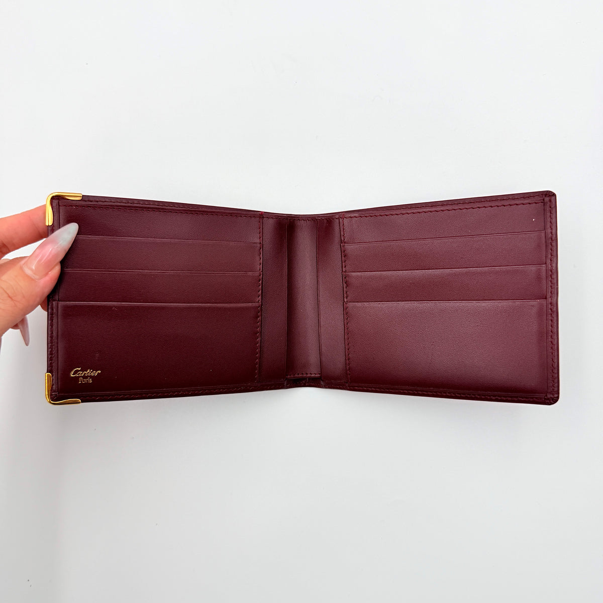 Cartier Must de Cartier burgundy leather bifold wallet with gold-tone corner accents shown against a white background.