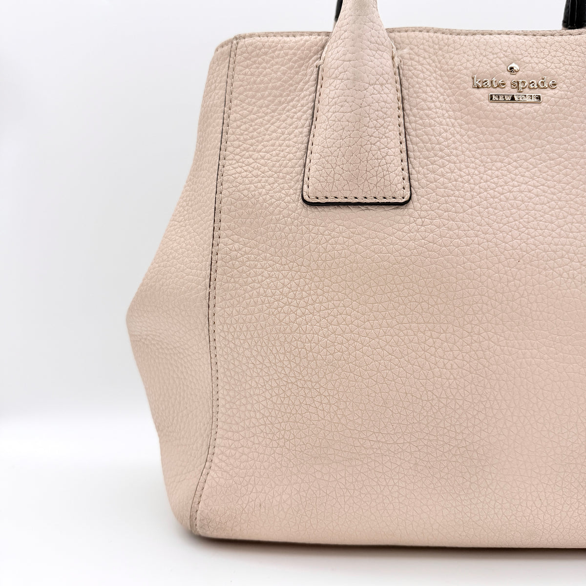 Kate Spade New York beige leather satchel with black handles, gold medallion, and grained texture against a white background.