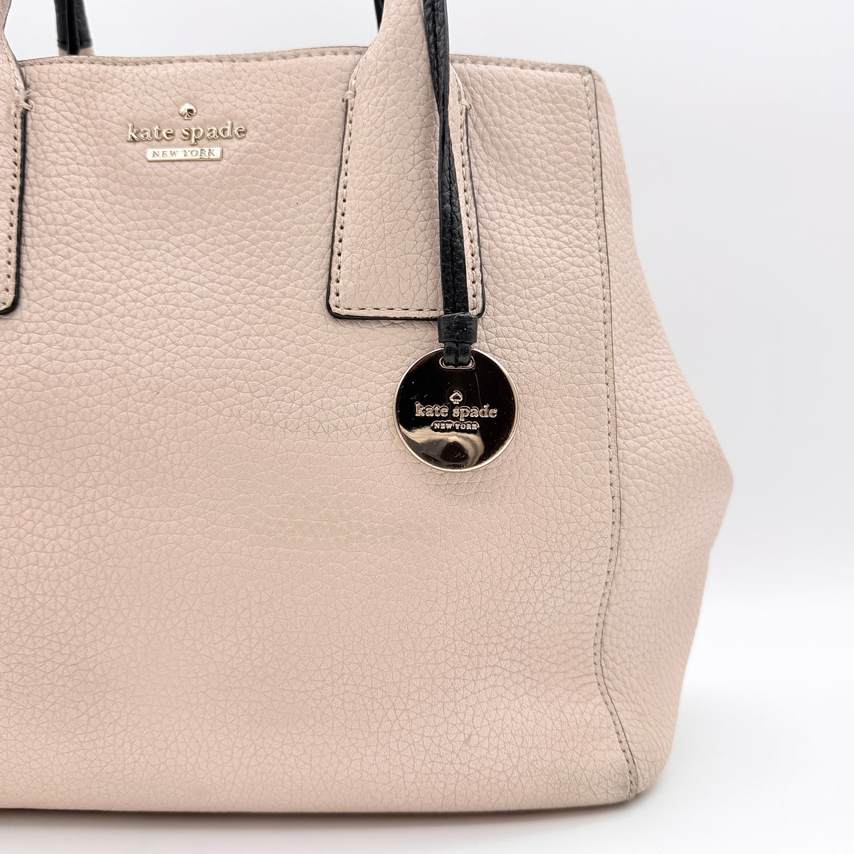 Kate Spade New York beige leather satchel with black handles, gold medallion, and grained texture against a white background.