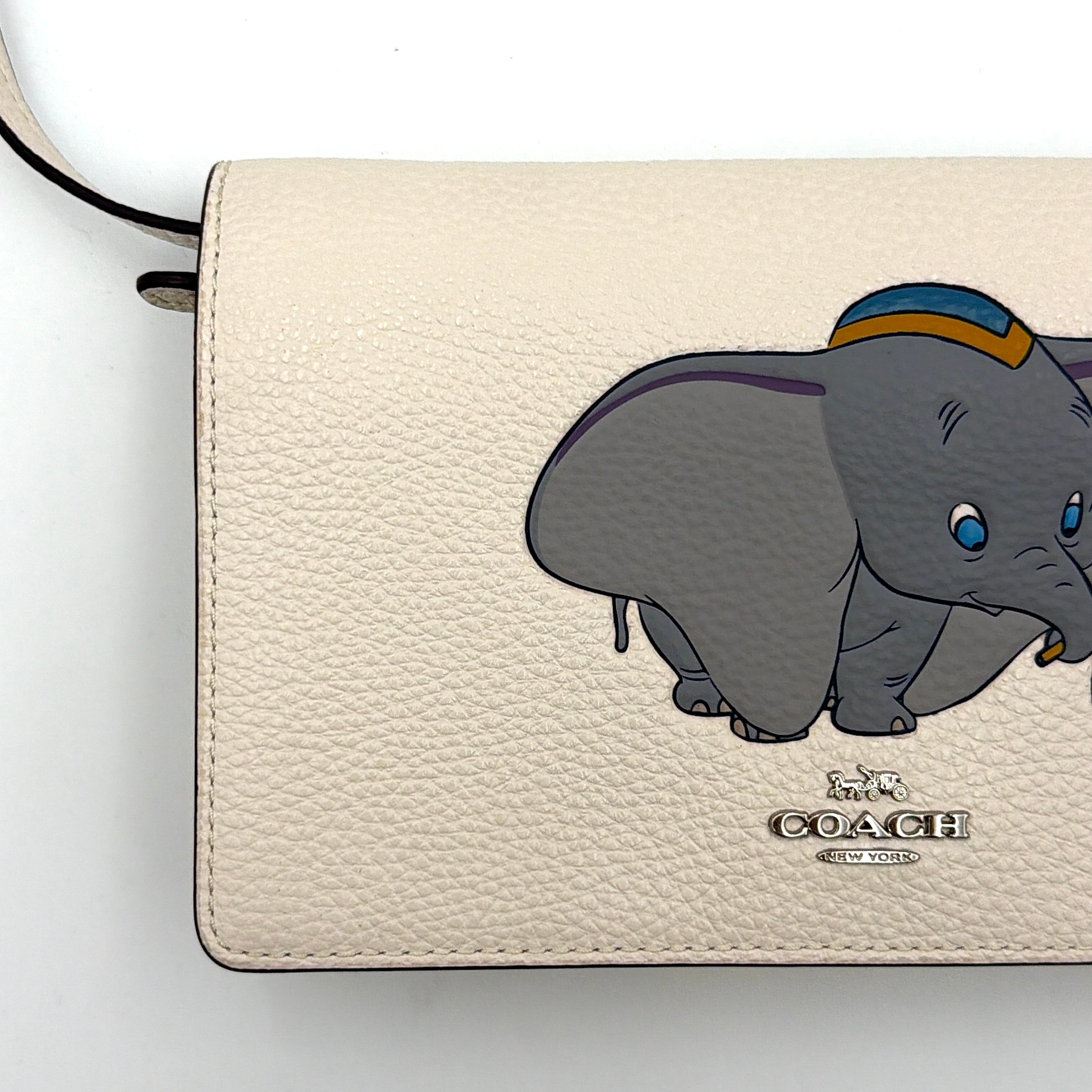 Coach x Disney Dumbo cream foldover crossbody bag with Dumbo illustration and slender strap, displayed against white background.
