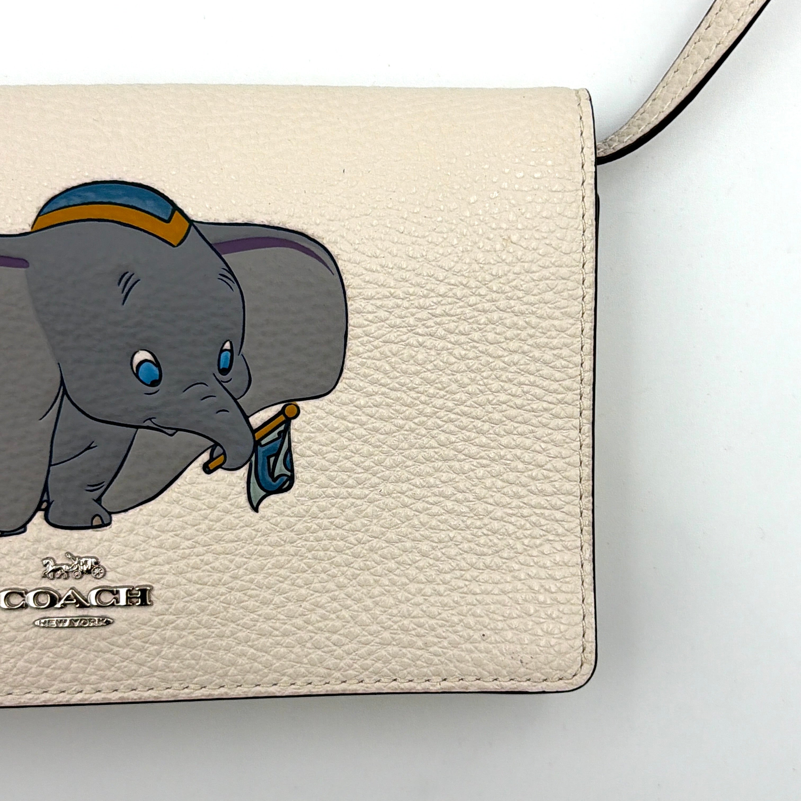 Coach x Disney Dumbo cream foldover crossbody bag with Dumbo illustration and slender strap, displayed against white background.