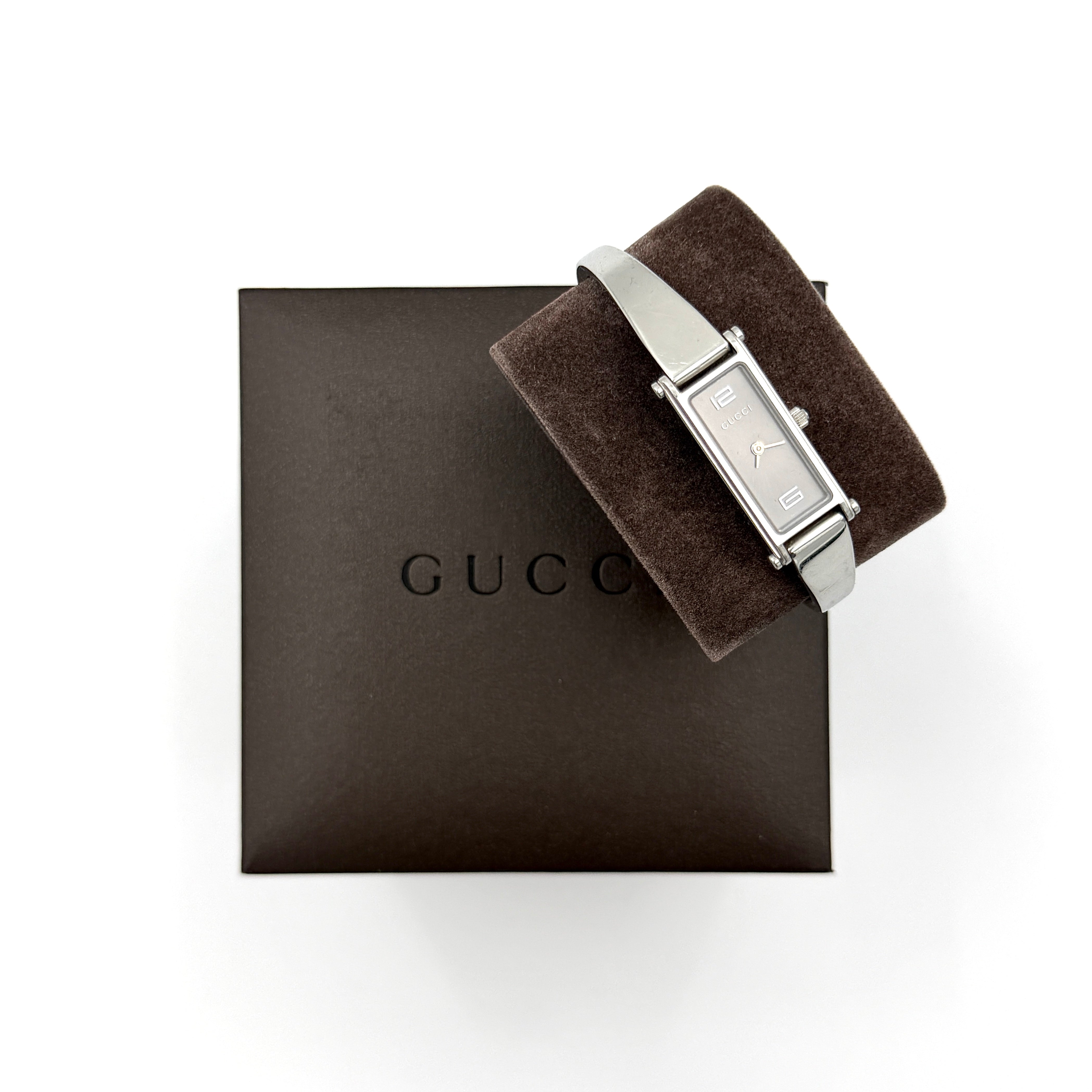 The Gucci 1500L Grey Dial Watch rests elegantly on its plush brown cushion atop a branded box, showcasing its sleek rectangular design and polished silver-tone bracelet.