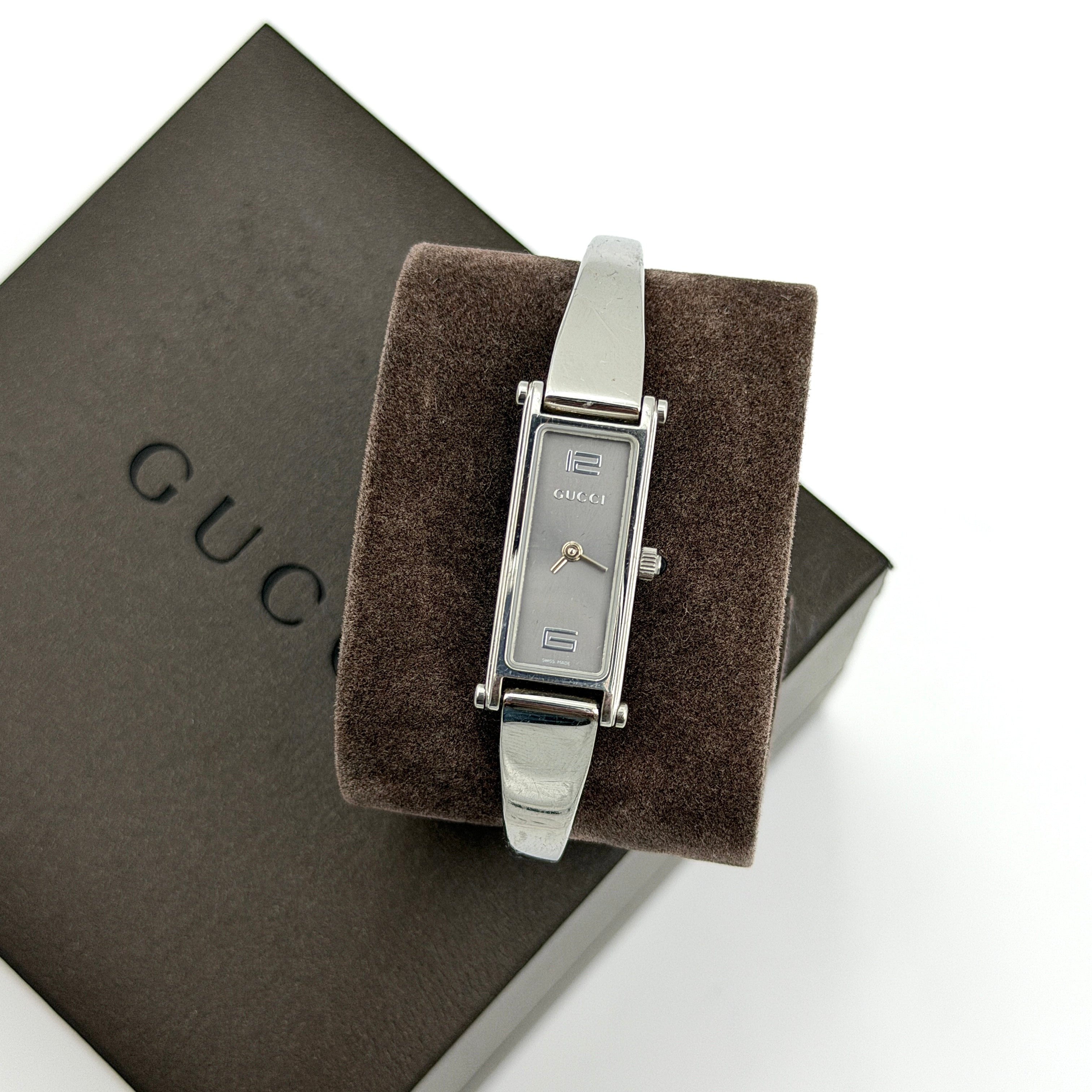 The Gucci 1500L Grey Dial Watch rests elegantly on its plush brown cushion atop a branded box, showcasing its sleek rectangular design and polished silver-tone bracelet.
