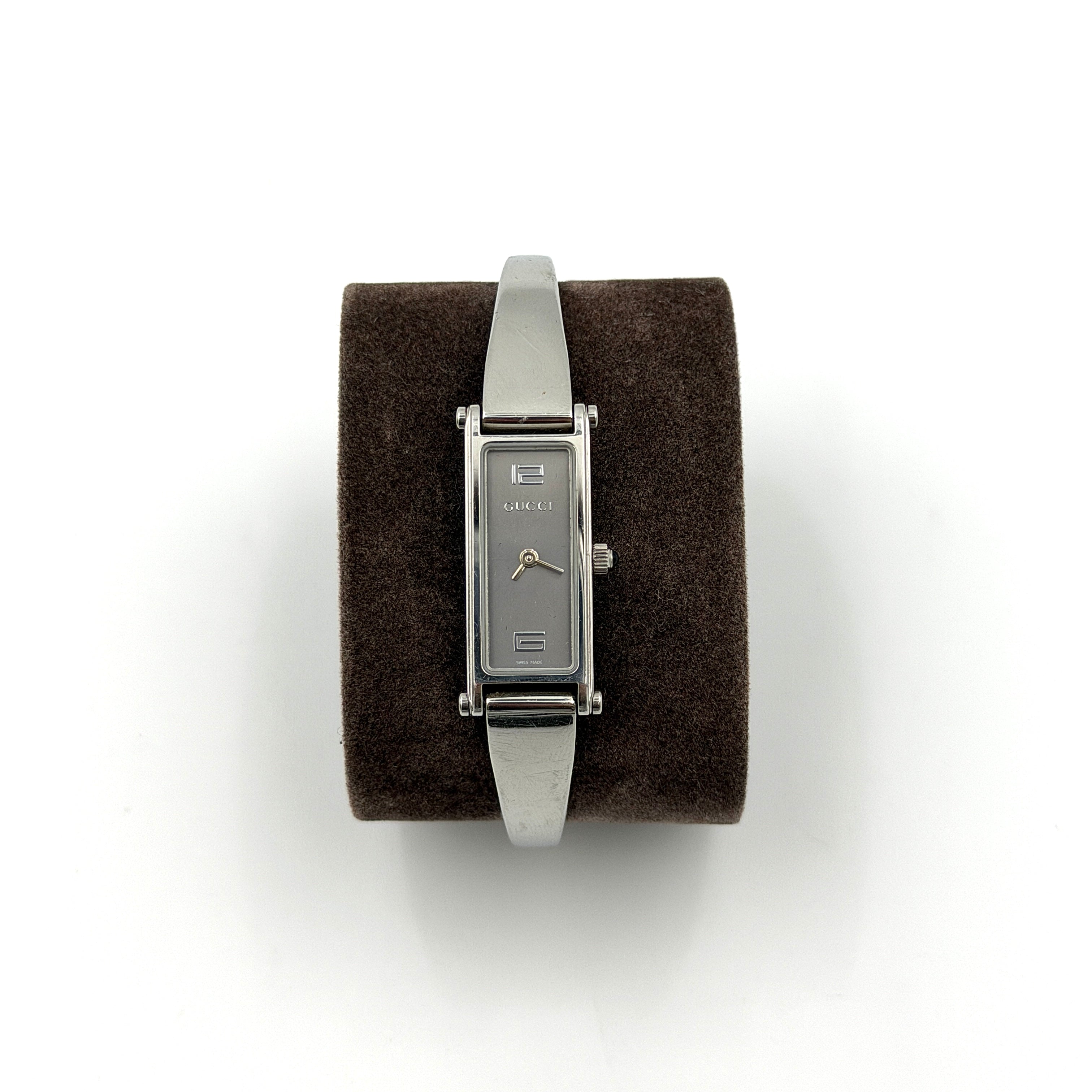 The Gucci 1500L Grey Dial Watch rests elegantly on its plush brown cushion atop a branded box, showcasing its sleek rectangular design and polished silver-tone bracelet.