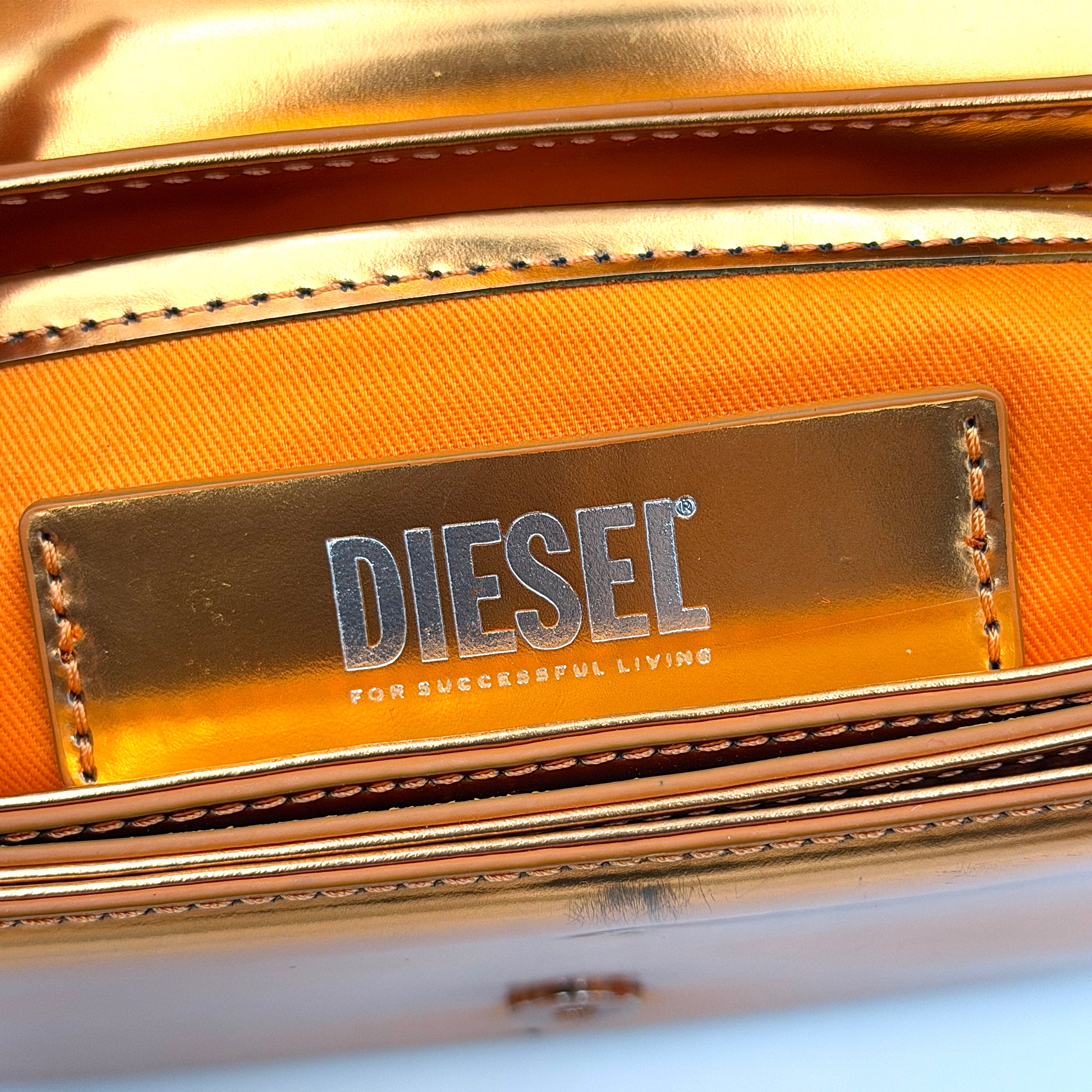 A Diesel 2Way 1DR Shoulder Bag in striking metallic gold with an embossed logo and detachable strap, displayed alongside its accessories.