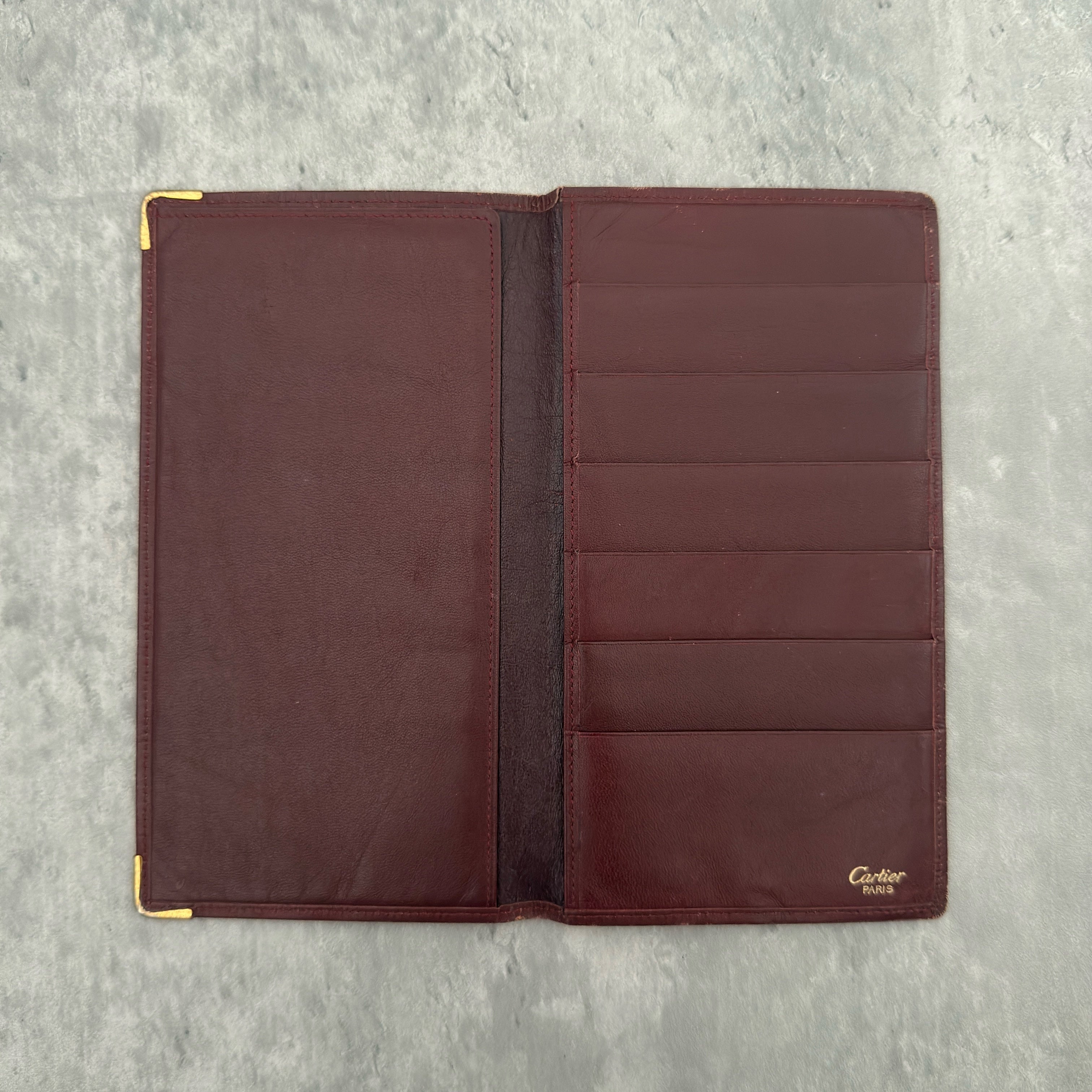 Cartier Must De Cartier Long Wallet in burgundy leather with embossed logo, gold-tone corner accents, and multiple compartments for organized storage.